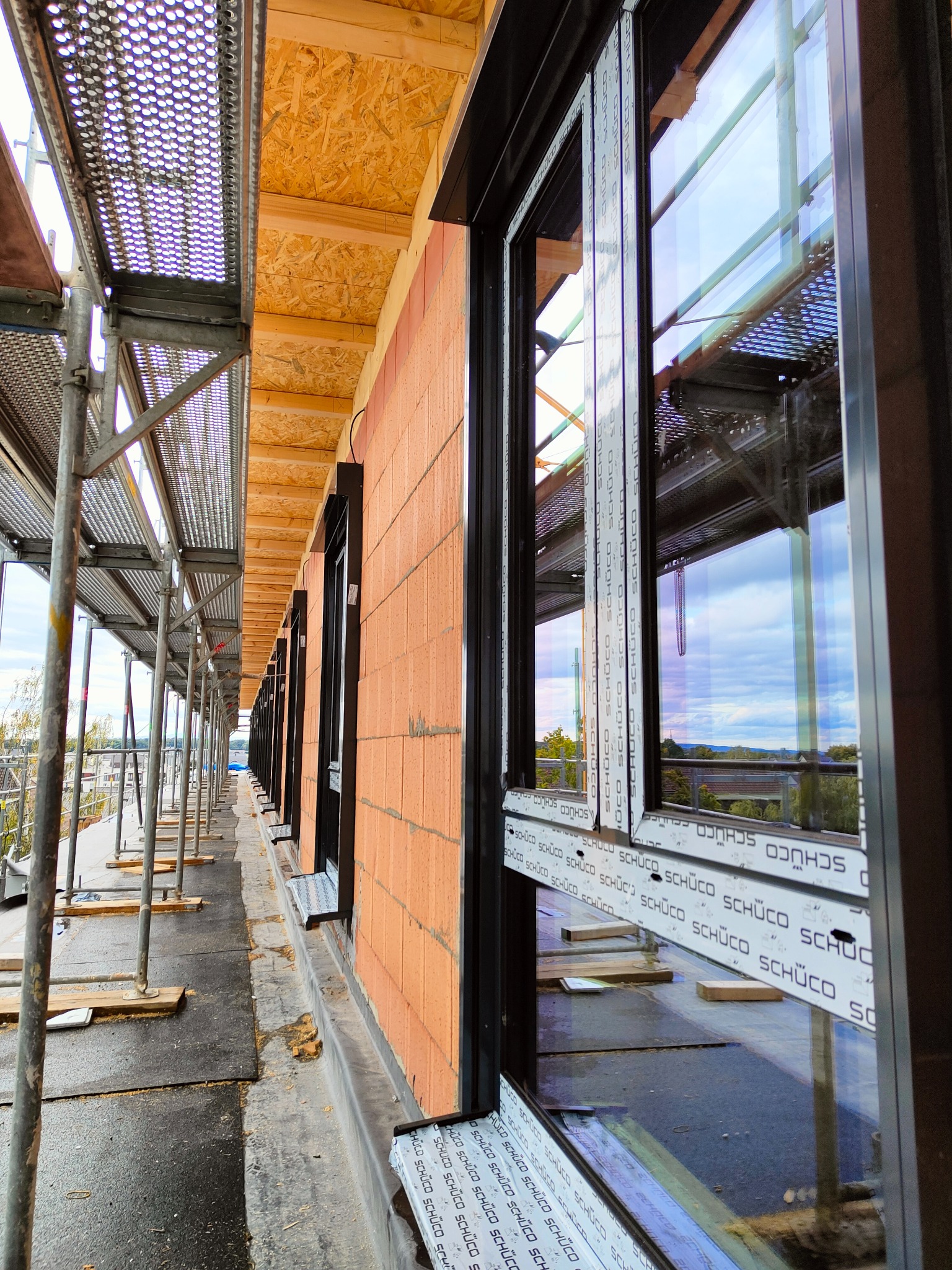 Montaż okien z czarnymi ramami na budynku z cegły, widoczne rusztowania i zabezpieczenia, okna marki Schuco.