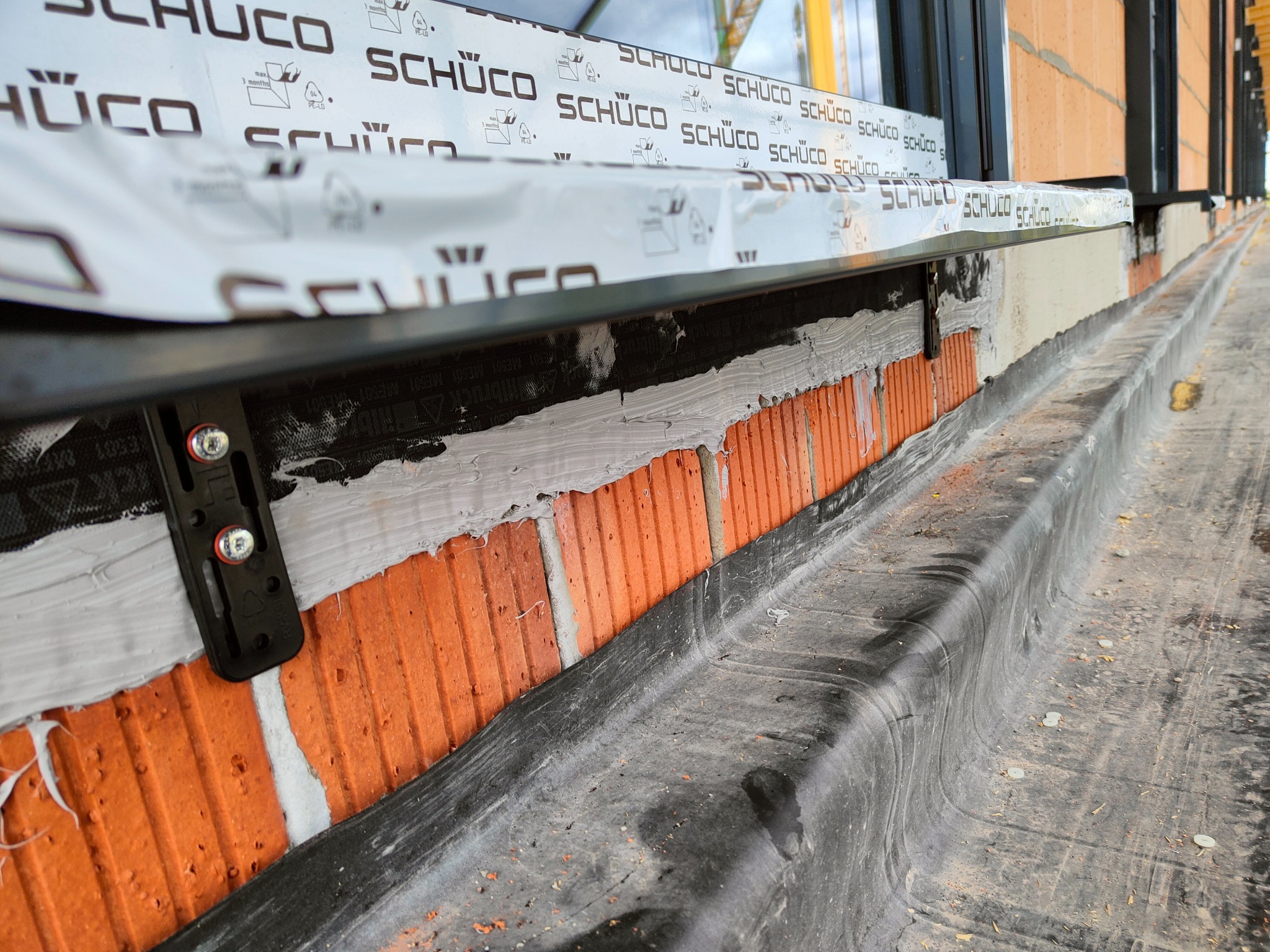 Montaż okna z widocznymi elementami mocującymi do muru z cegły oraz folią ochronną producenta na ramie okiennej.