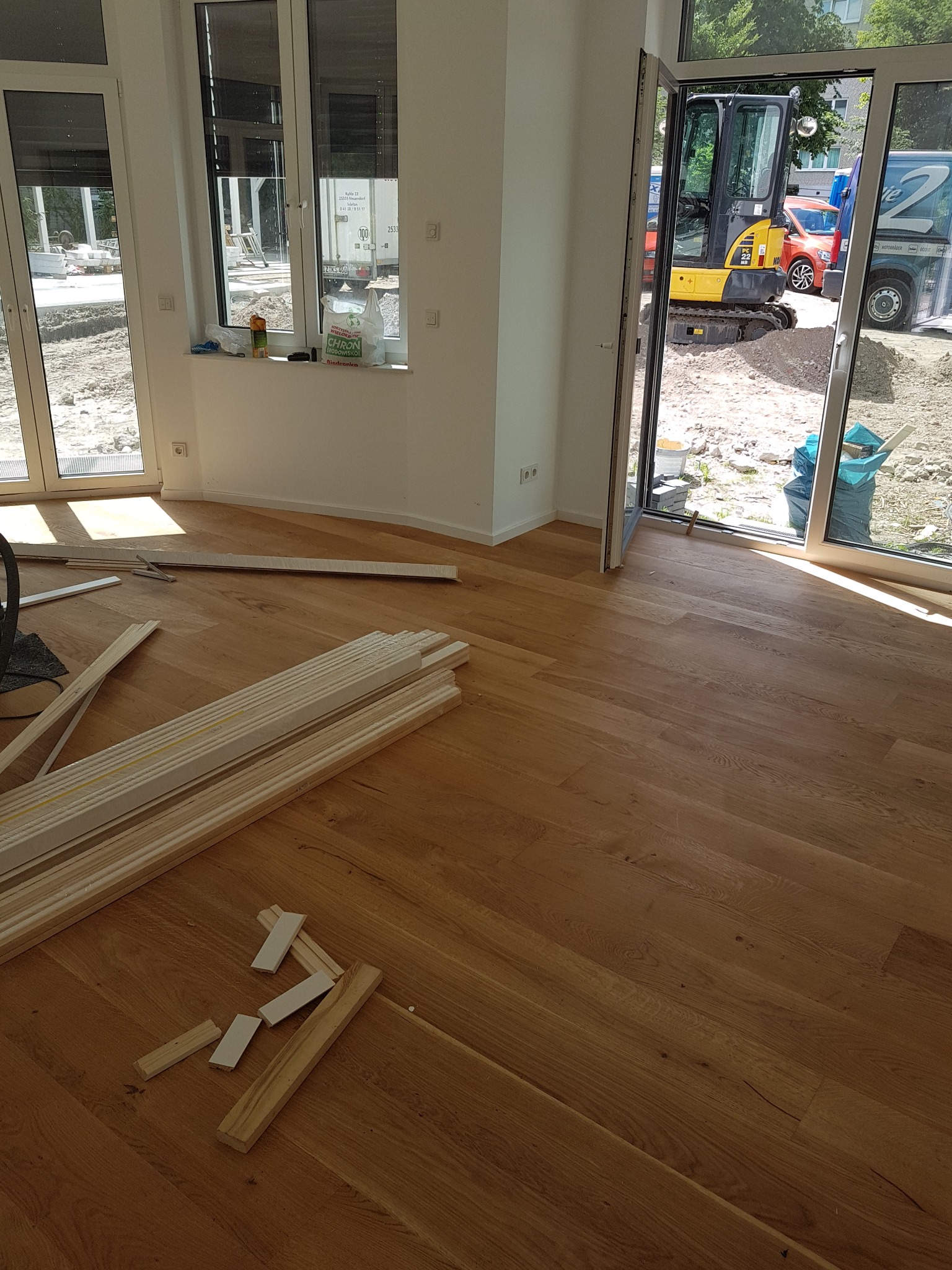 Wnętrze w trakcie prac wykończeniowych: ułożona drewniana podłoga, listwy przypodłogowe w paczkach, widok na teren budowy z mini koparką przez otwarte okno.