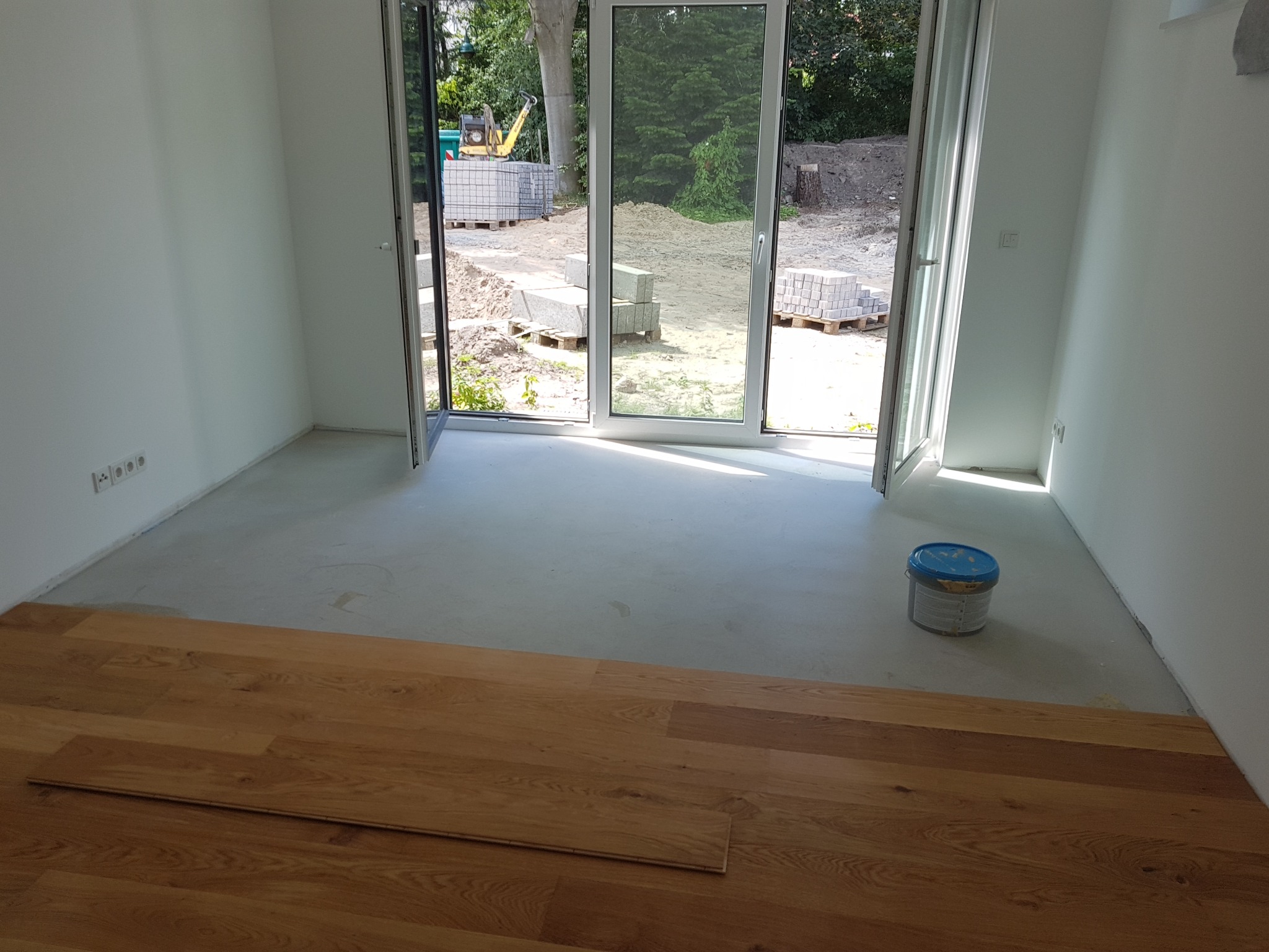 Wnętrze pokoju w trakcie wykańczania: fragment ułożonej drewnianej podłogi, ściany pomalowane na biało, otwarte drzwi balkonowe z widokiem na teren budowy z kostką brukową i mini koparką.