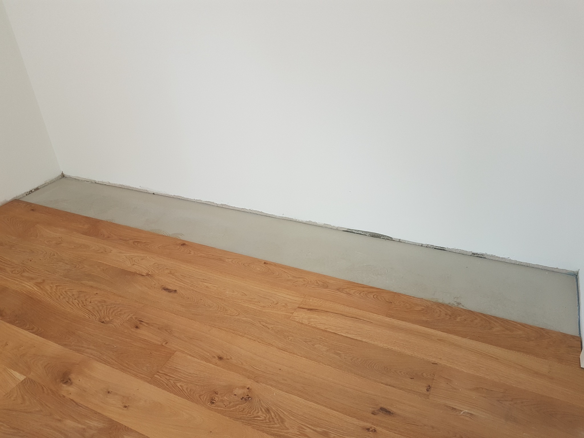 Fragment układanej podłogi drewnianej w rogu pomieszczenia, widoczny surowy betonowy podkład przy ścianie.
