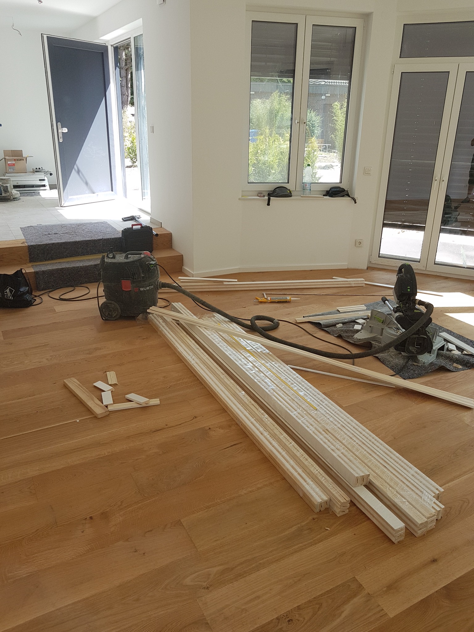 Przygotowane listwy przypodłogowe, odkurzacz Wurth, piła elektryczna i akcesoria rozłożone na drewnianej podłodze w jasnym wnętrzu z dużymi oknami i otwartymi drzwiami.