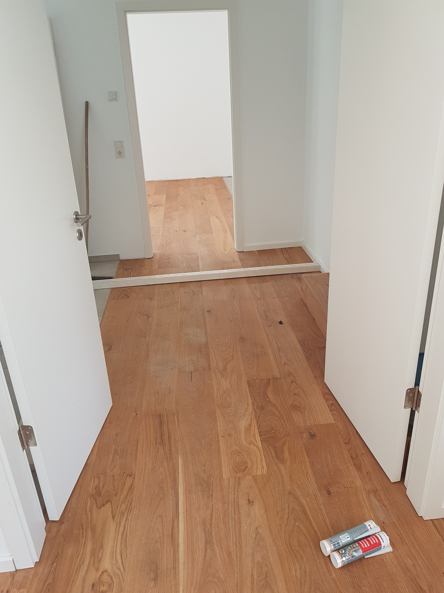 Montaż drewnianej podłogi w korytarzu, widoczne białe drzwi i listwa progowa, dwie tuby kleju uszczelniającego leżą na podłodze.