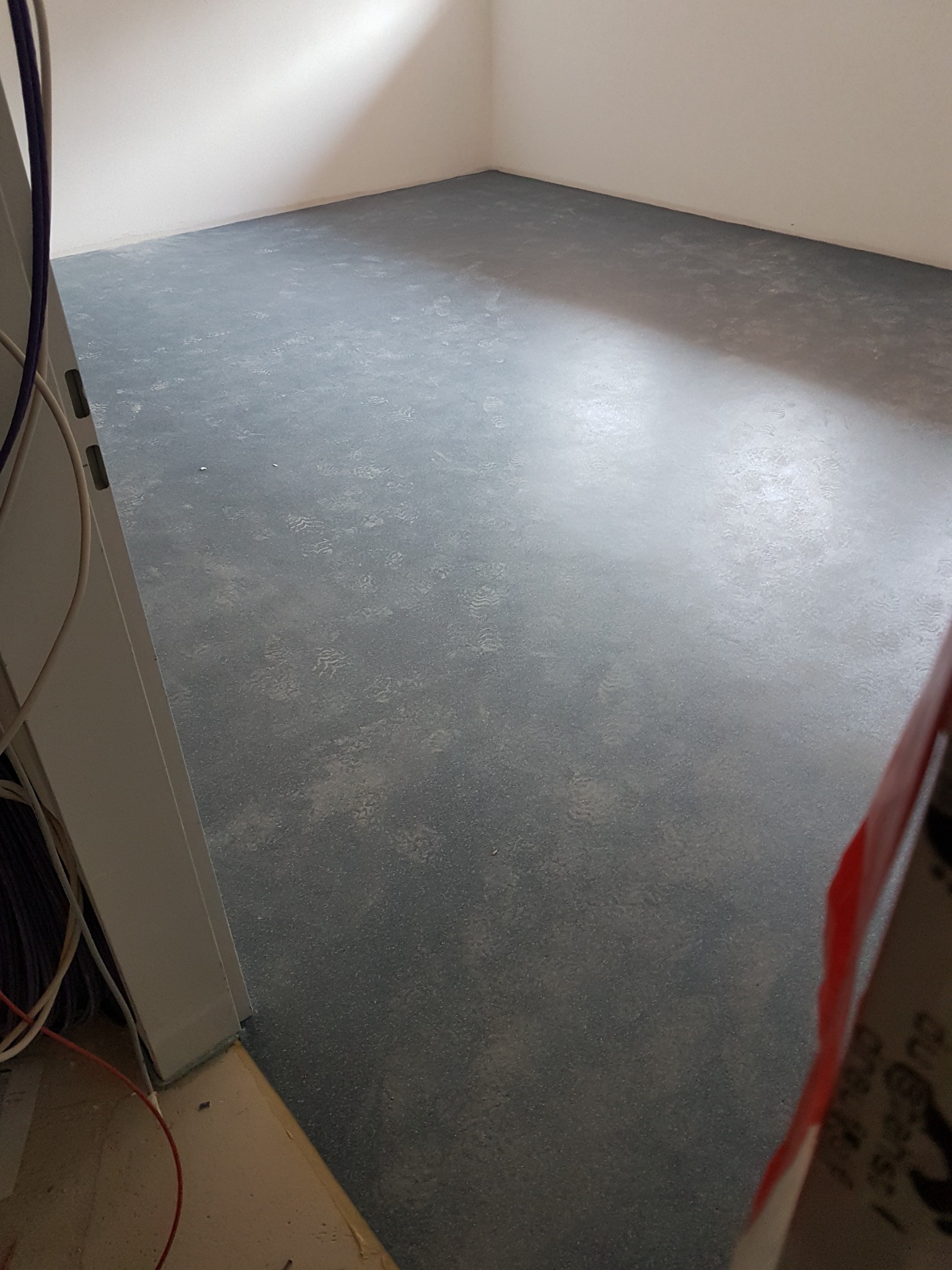 Szare, wzorzyste wykończenie podłogi w pustym pomieszczeniu z białymi ścianami, widoczne kable i fragment futryny drzwi.