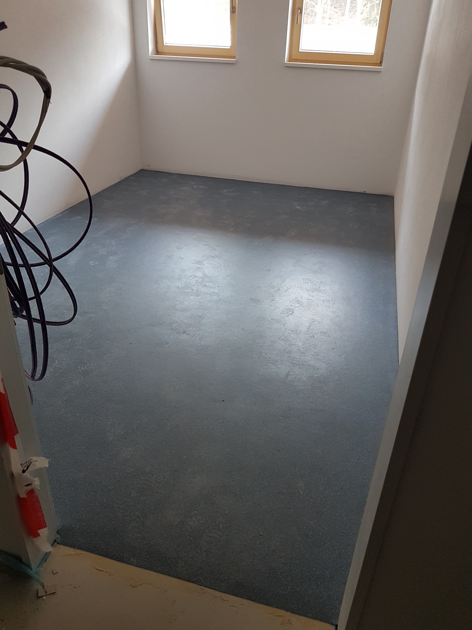 Pokój po wylewce posadzki w odcieniach szarości, z widocznymi przewodami elektrycznymi w rogu i dwoma oknami z drewnianą ramą.