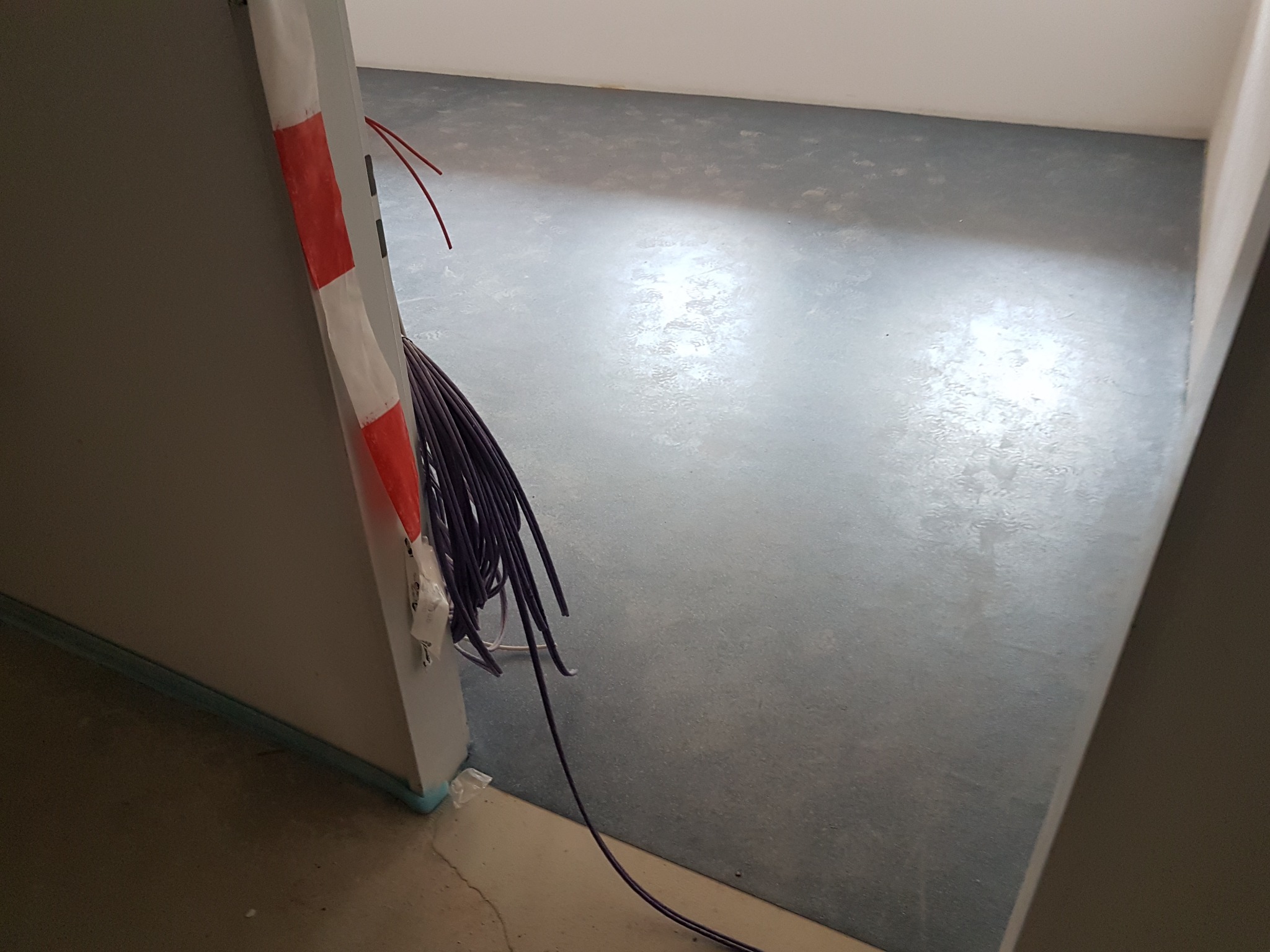 Szary, gładki wylewka podłogowa w trakcie schnięcia, widoczne kable elektryczne przy ścianie, zabezpieczone taśmą ostrzegawczą.
