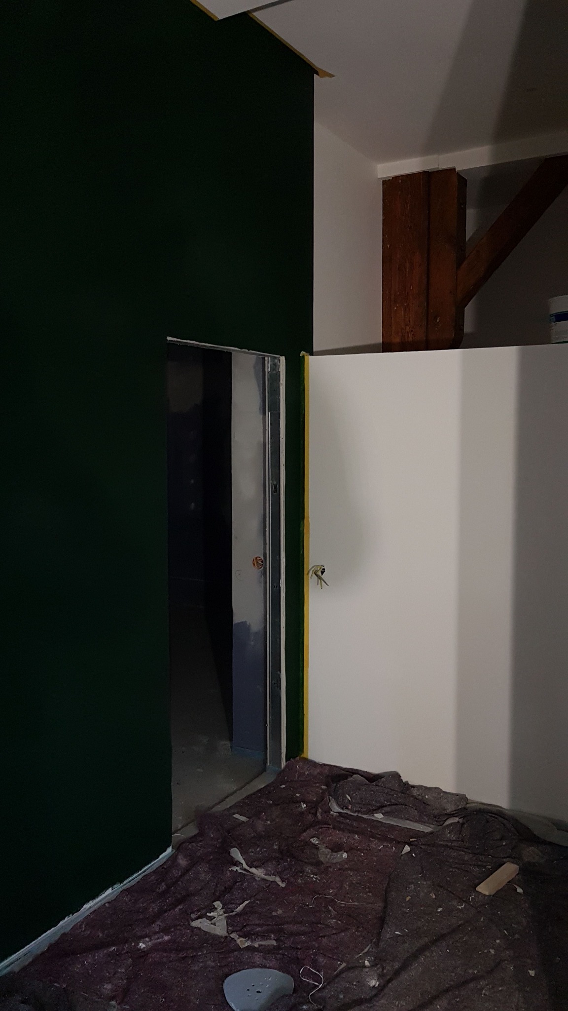 Wnętrze pokoju w trakcie malowania: ściana pomalowana na ciemnozielono, druga ściana biała, zabezpieczona taśmą malarską. Na podłodze rozłożona plandeka malarska. Widoczny fragment drewnianej belki...