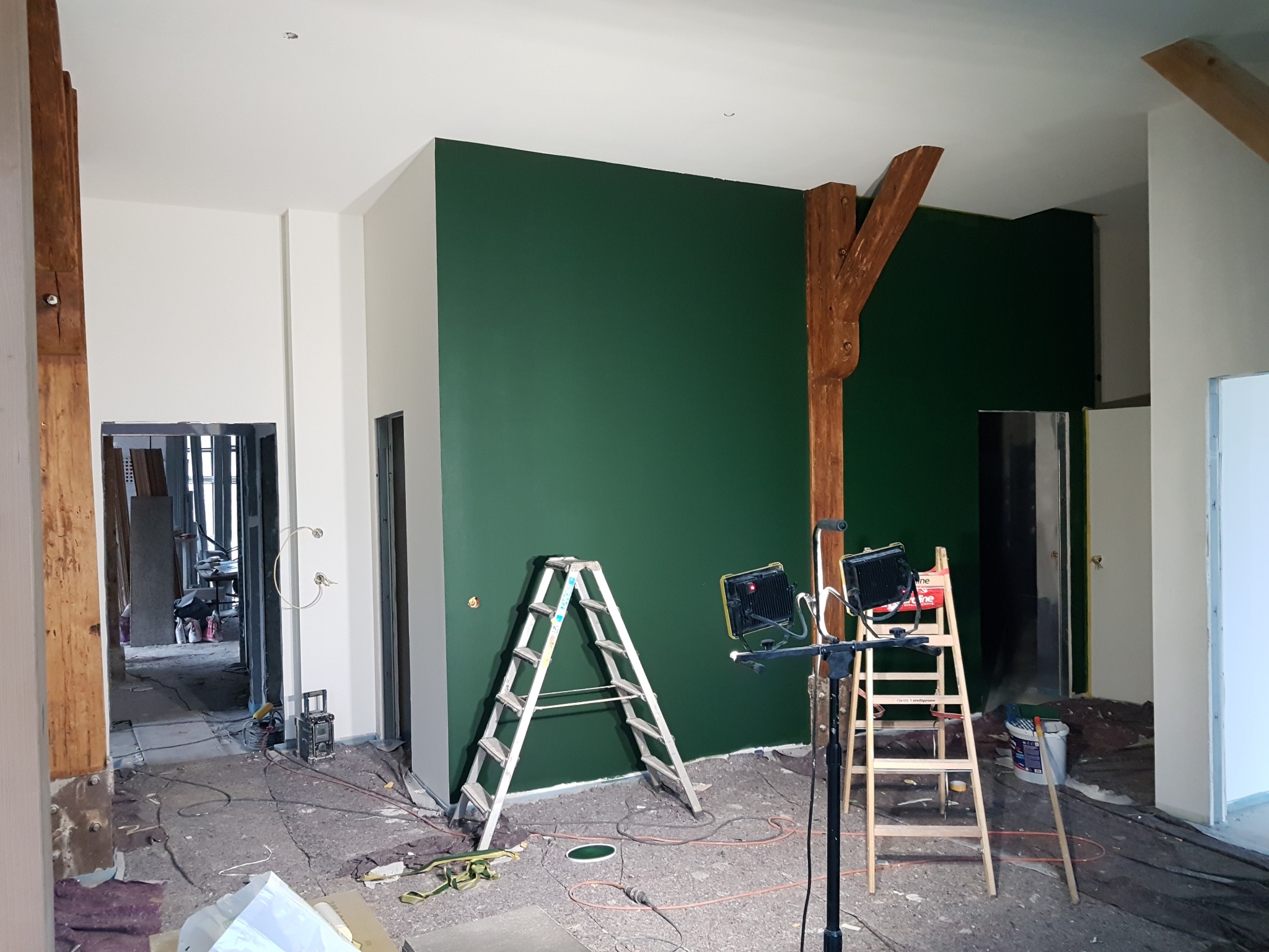 Wnętrze w trakcie malowania: ściana pomalowana na ciemnozielono, drewniane belki konstrukcyjne, dwie drabiny (aluminiowa i drewniana), lampy budowlane, widoczne przewody i narzędzia malarskie...