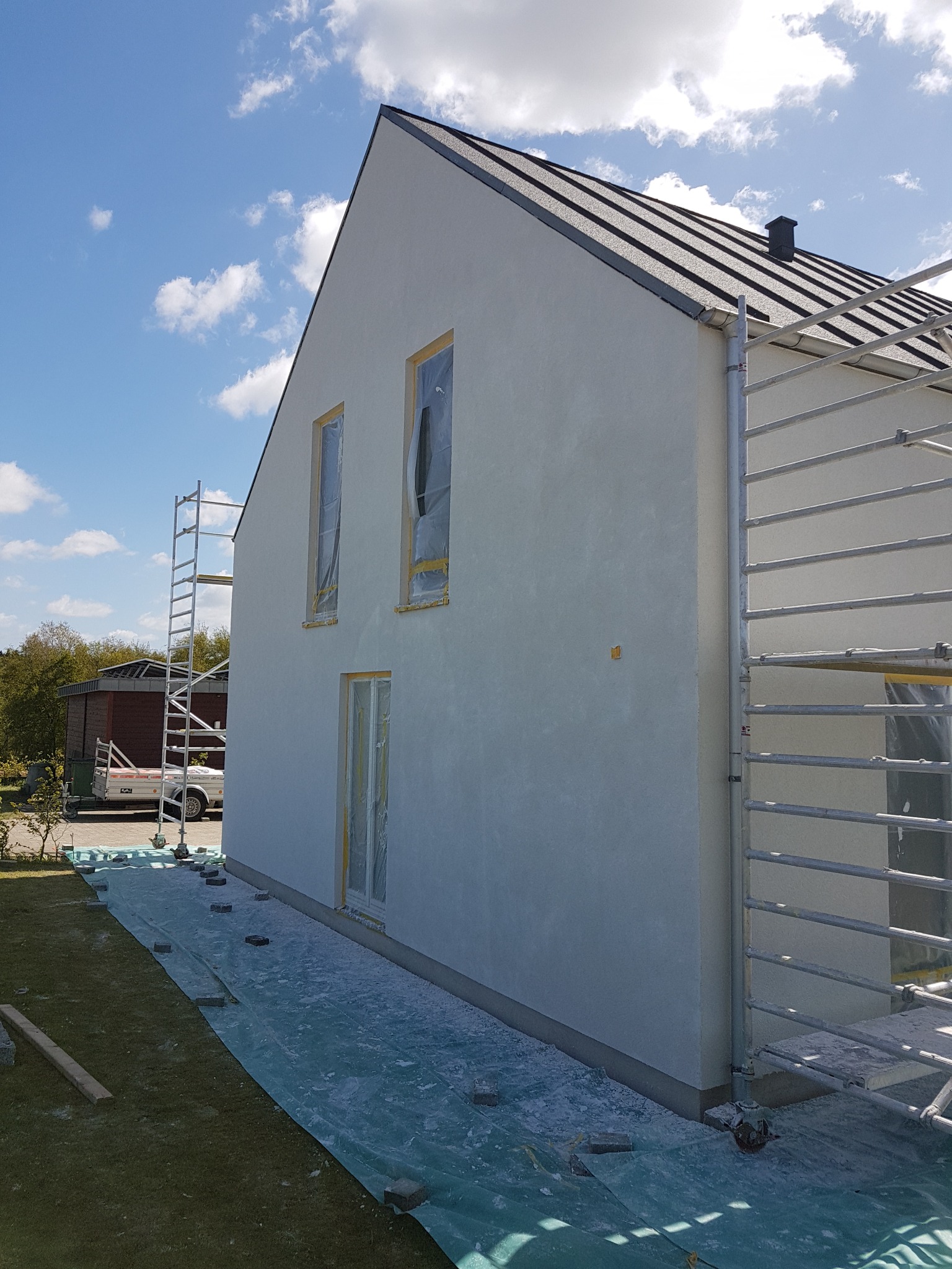 Boczny widok na dom jednorodzinny w trakcie ocieplania, z widocznymi rusztowaniami, zabezpieczonymi oknami folią i rozłożoną folią ochronną na trawniku.