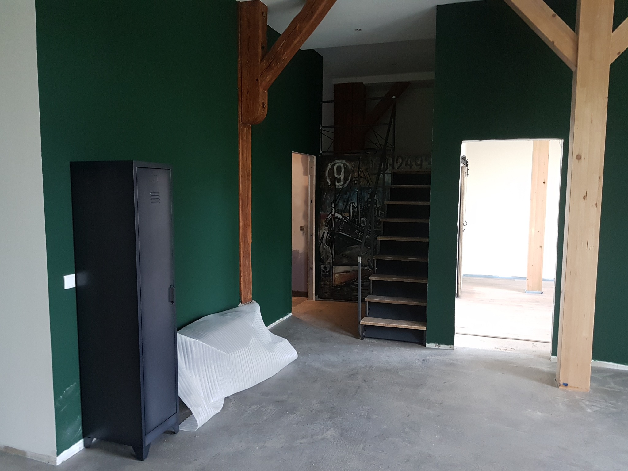 Wnętrze w trakcie remontu: butelkowa zieleń ścian, surowe drewniane belki konstrukcyjne, metalowa szafka i zabezpieczone folią przedmioty na szarym betonowym podłożu, schody z metalową konstrukcją...