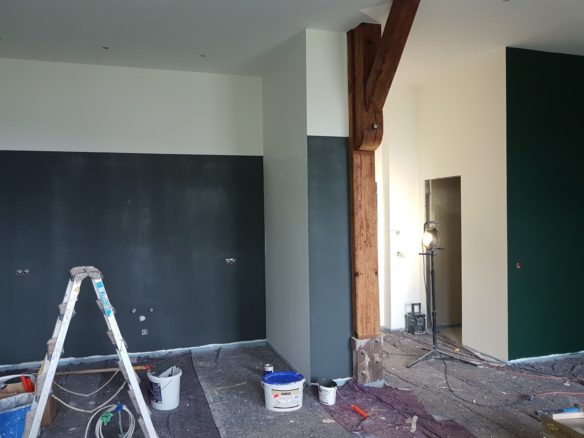 Wnętrze w trakcie malowania we Włocławku: ściany w dwóch odcieniach szarości i bieli, drewniany filar, drabina malarska, puszki z farbą i lampa oświetlająca pomieszczenie.