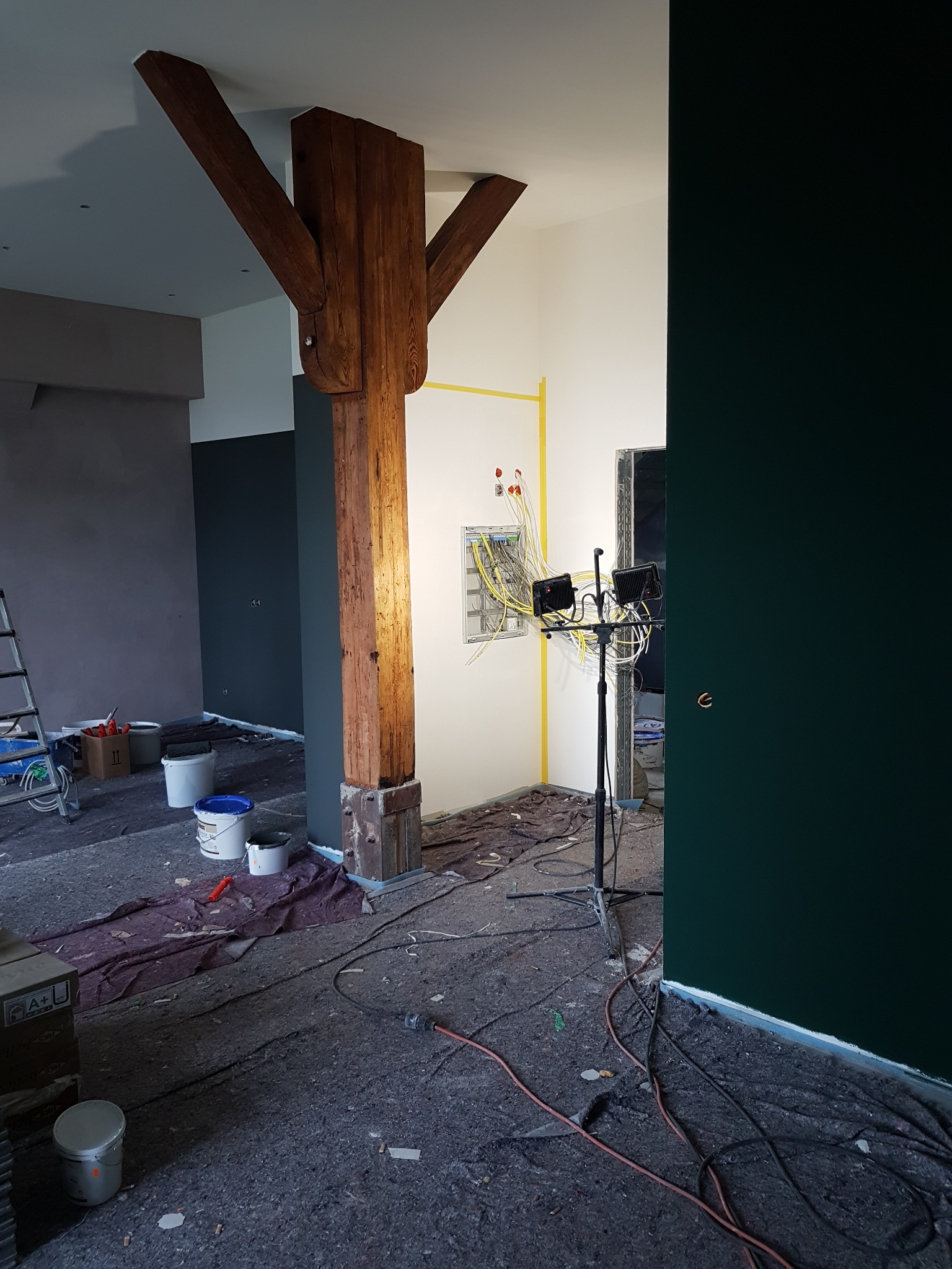 Wnętrze w trakcie remontu: widoczna drewniana kolumna wspierająca strop, ściany przygotowane do malowania z widoczną instalacją elektryczną oraz profesjonalne oświetlenie budowlane.