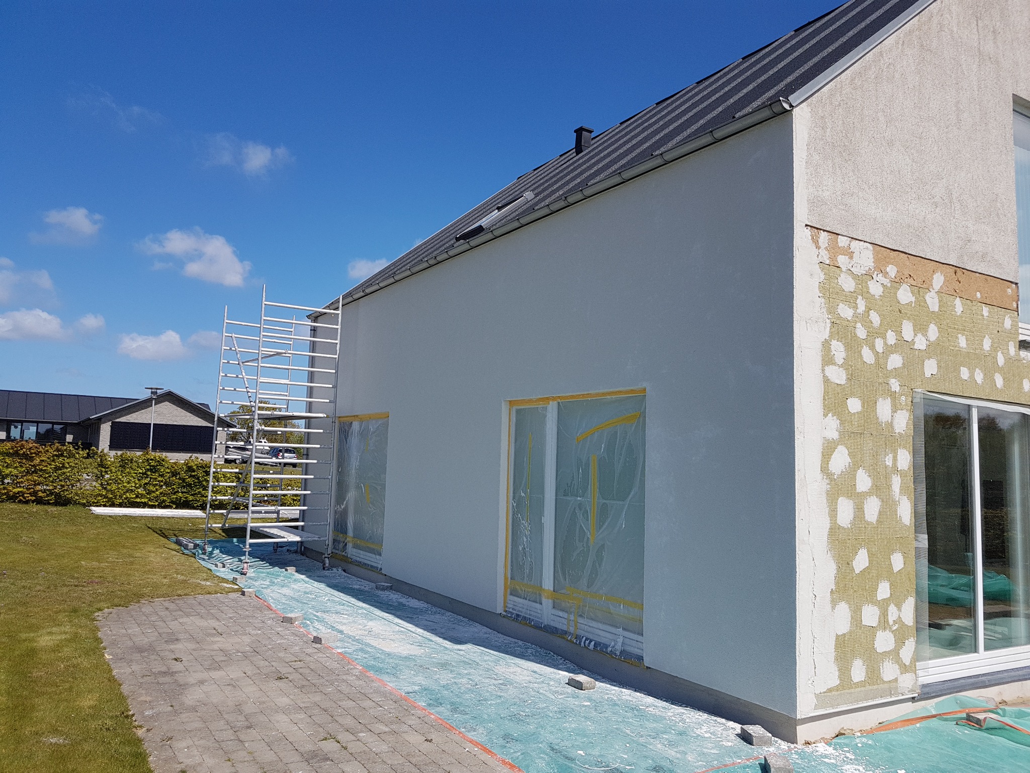 Dom jednorodzinny w trakcie ocieplania, widoczna warstwa izolacji termicznej z fragmentami nałożonego kleju, okna zabezpieczone folią malarską, rusztowanie aluminiowe przy ścianie.