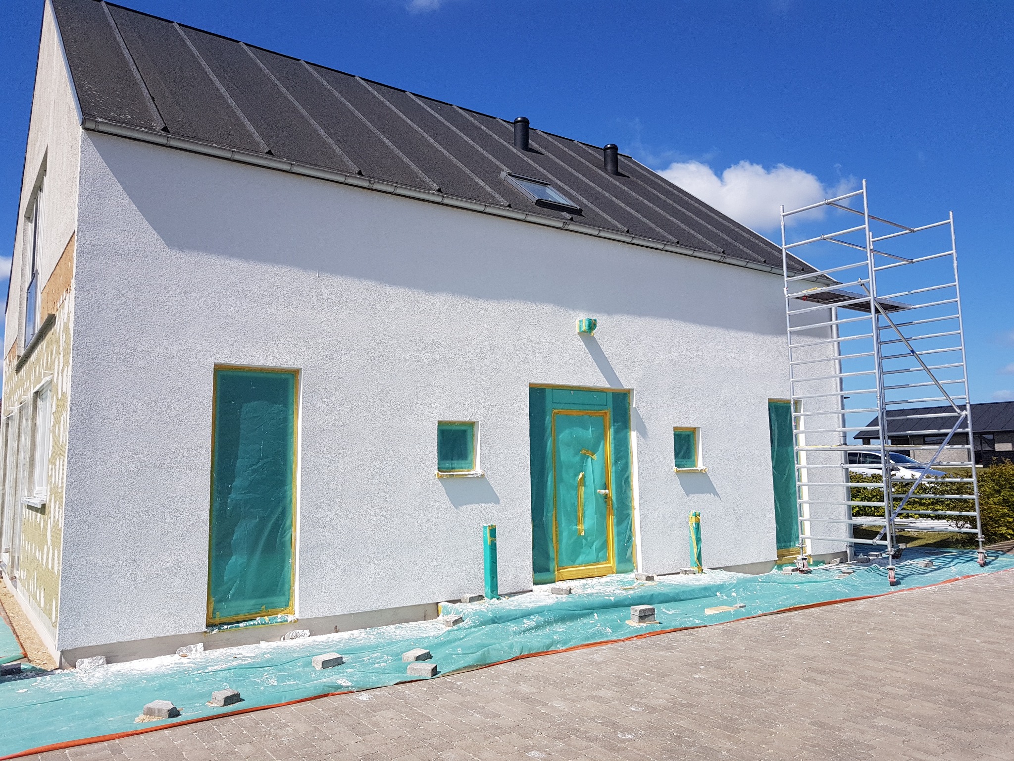 Biały dom w trakcie ocieplania, z widocznym rusztowaniem, zabezpieczonymi folią oknami i drzwiami, oraz fragmentem ocieplonej ściany z widoczną warstwą izolacyjną.