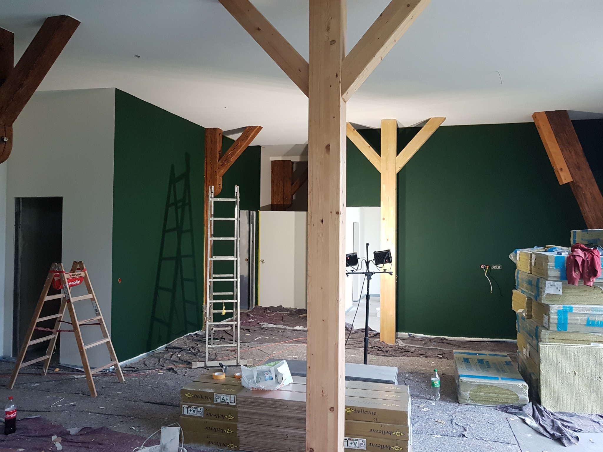 Wnętrze w trakcie malowania na ciemnozielono, widoczne drewniane belki konstrukcyjne, drabiny, reflektory, materiały budowlane i zabezpieczenia podłogi.