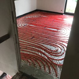 Domagalski instalacje - Czerwone rury ogrzewania podłogowego ułożone spiralnie na siatce w surowym wnętrzu poddasza, widoczne fragmenty ściany z oknem i niezakończone obramowanie przejścia.