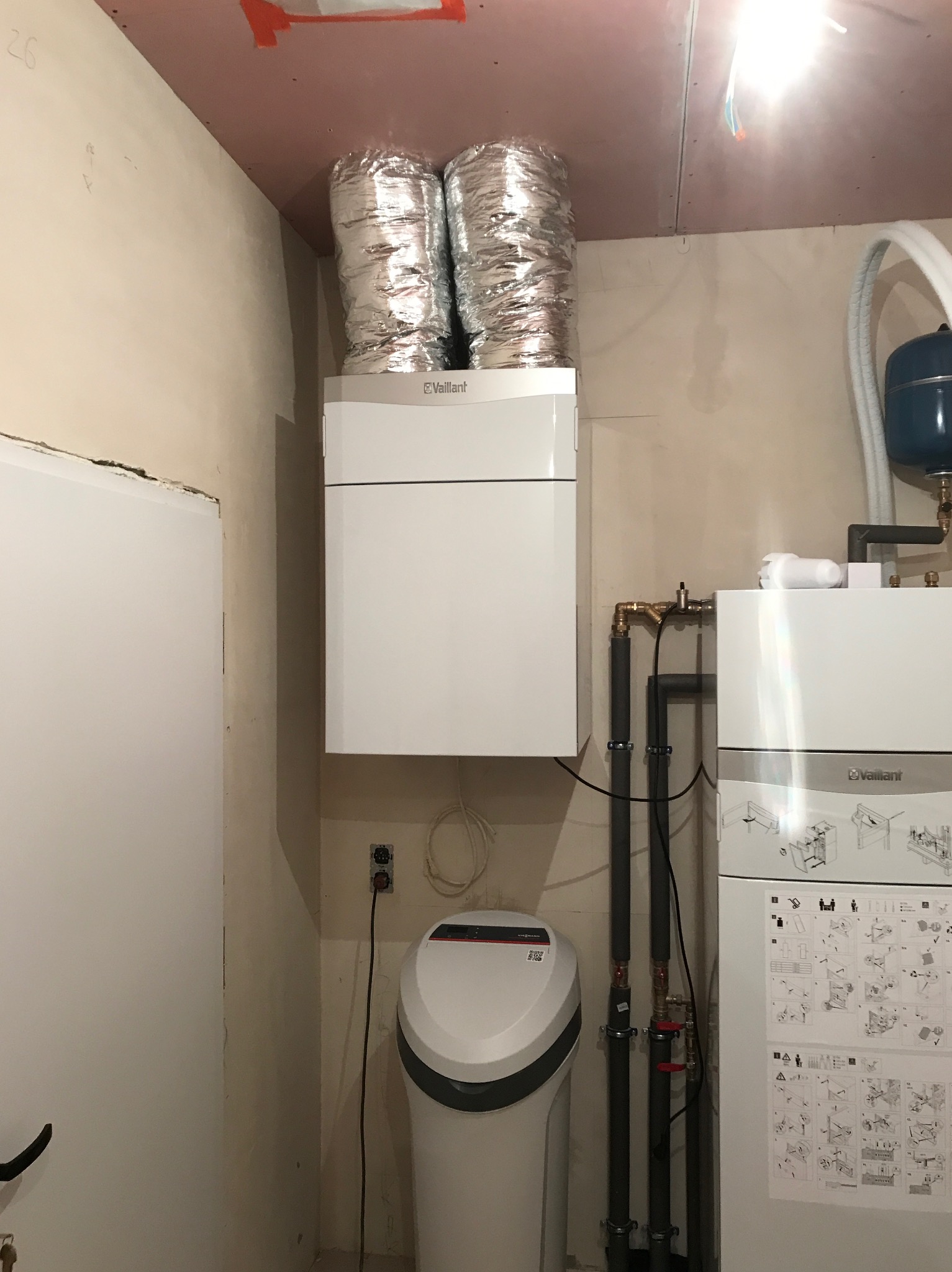Biała jednostka rekuperacji marki Vaillant z aluminiowymi przewodami wentylacyjnymi na suficie, obok instalacja hydrauliczna i zmiękczacz wody, widoczne rury i zawory, ściany w stanie surowym.