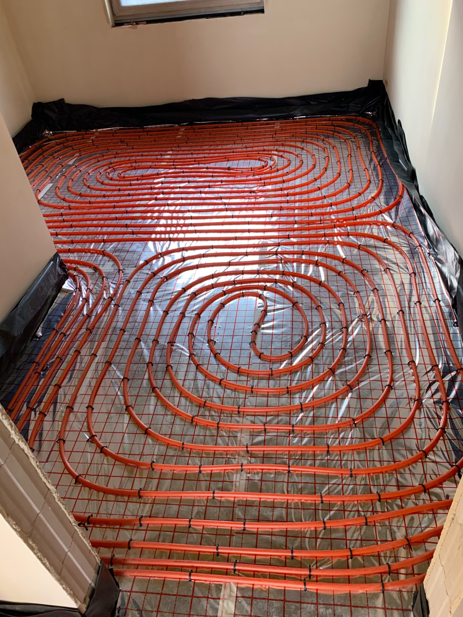 Pomarańczowe rury ogrzewania podłogowego ułożone spiralnie na srebrnej folii izolacyjnej, przymocowane czarnymi klipsami do siatki, widok z góry w jasnym pomieszczeniu.