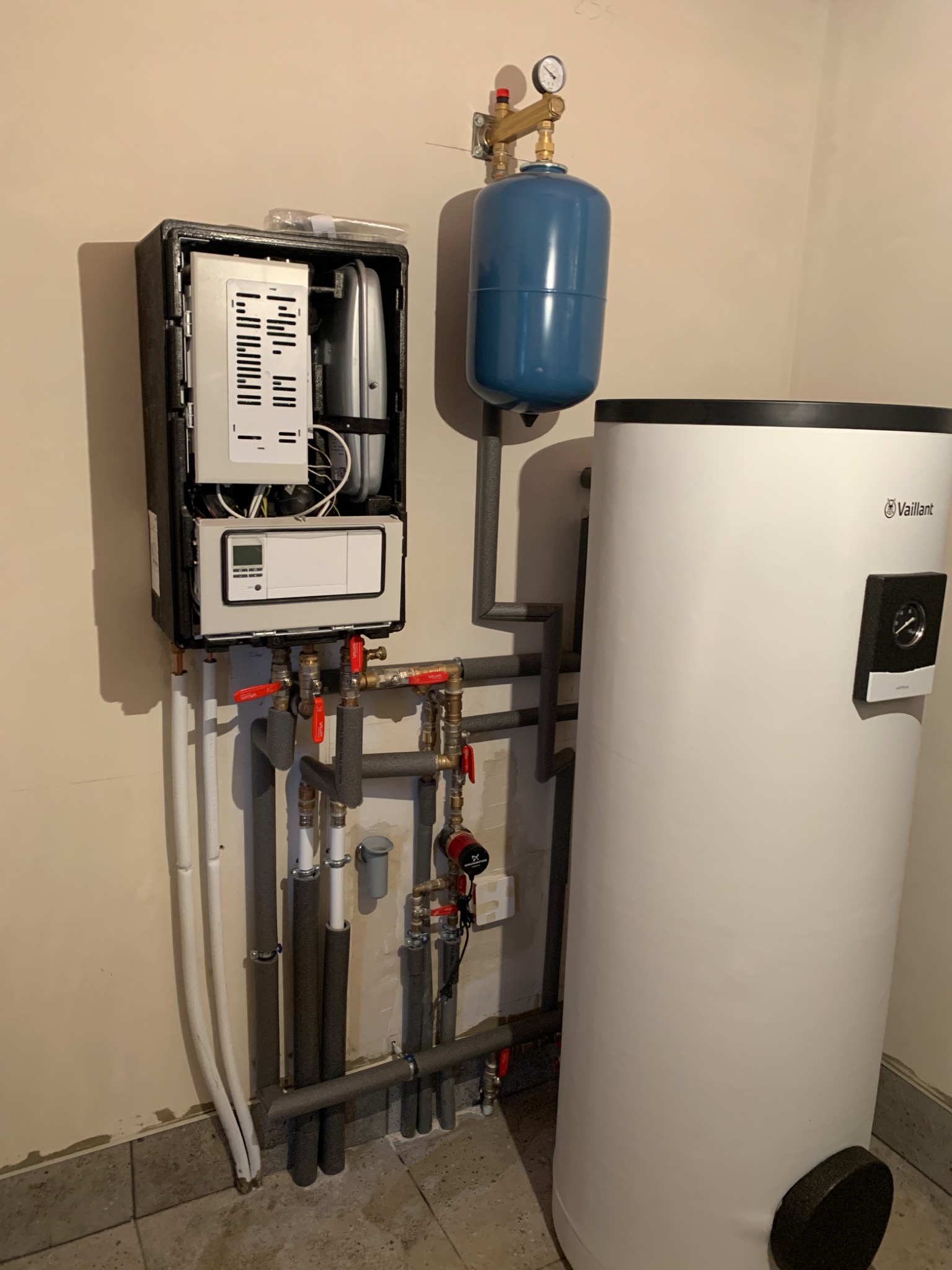 Otwarty panel kotła grzewczego Vaillant z widocznymi elementami wewnętrznymi, podłączony do pionowego, białego zbiornika i niebieskiego naczynia wzbiorczego, z widocznymi rurami i zaworami.