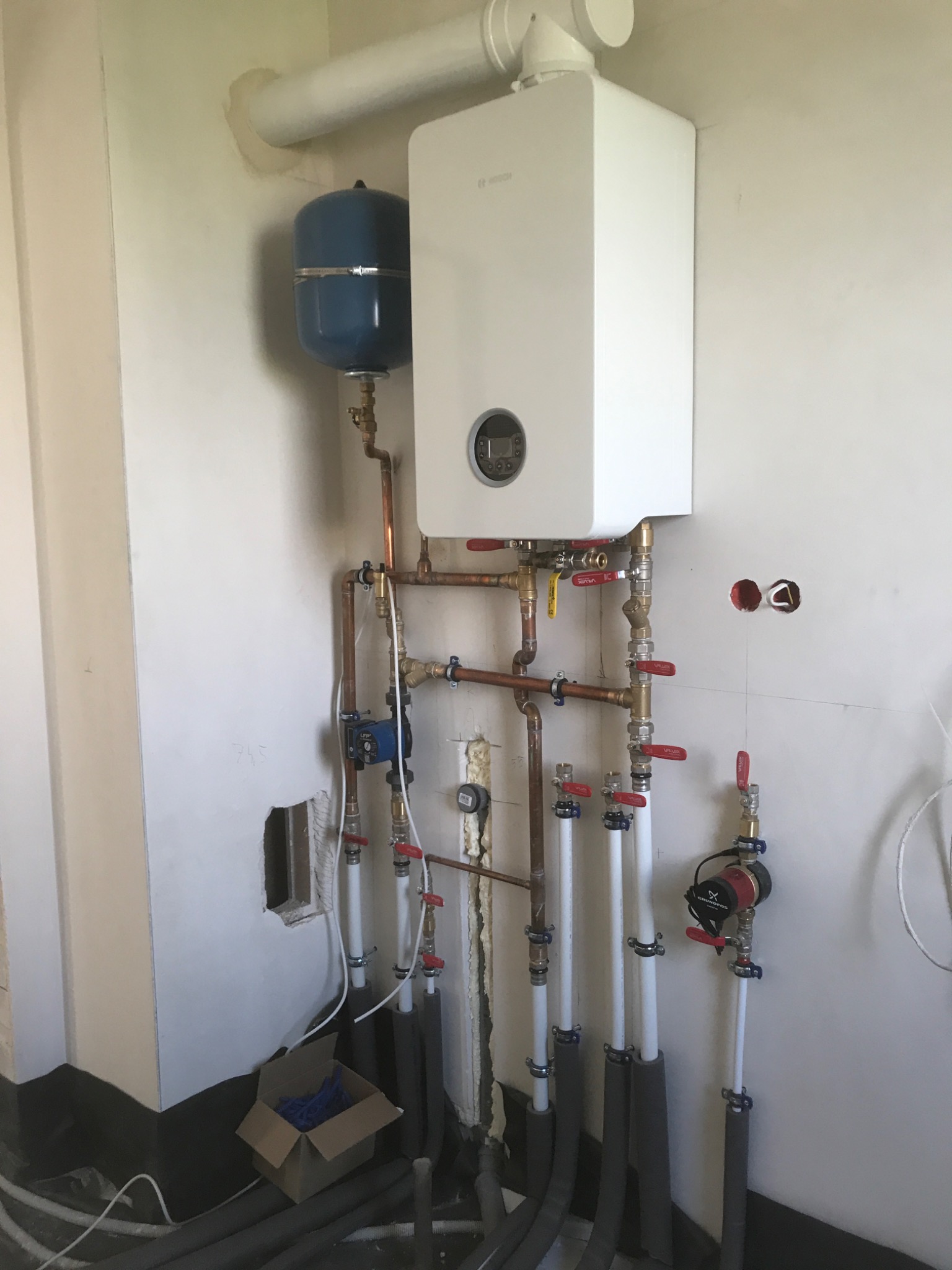 Biały wiszący piec grzewczy Bosch z niebieskim naczyniem przeponowym, połączony rurami miedzianymi i białymi rurami PCV z czerwonymi zaworami, widoczne otwory w ścianie i kartonowe pudełko...