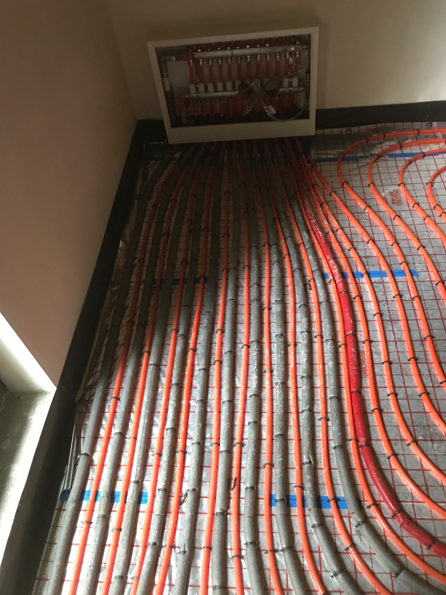 Widok z góry na rozłożone rury ogrzewania podłogowego, częściowo zalane szlichtą, z rozdzielaczem w białej obudowie zamontowanym na ścianie.