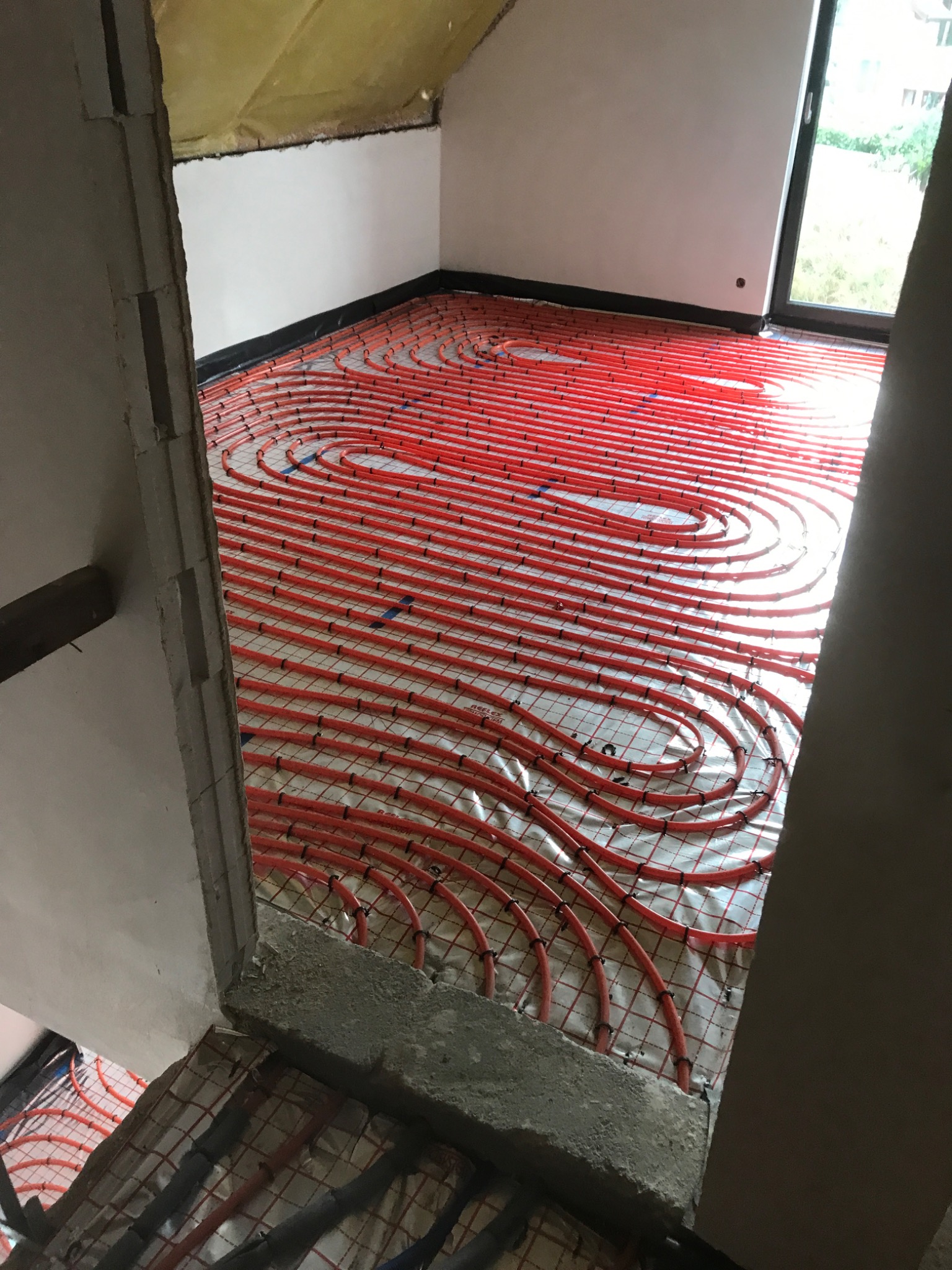 Czerwone rury ogrzewania podłogowego ułożone spiralnie na siatce w surowym wnętrzu poddasza, widoczne fragmenty ściany z oknem i niezakończone obramowanie przejścia.