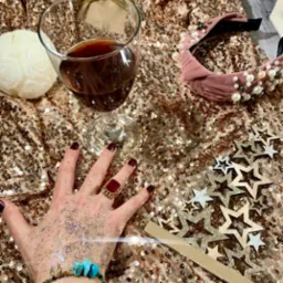 Kieliszek czerwonego wina na złotym, cekinowym tle obok ręki z brokatem, pierścionkiem z czerwonym kamieniem i bransoletką z turkusowymi kamieniami, różowa opaska z perłami, drewniane gwiazdki...