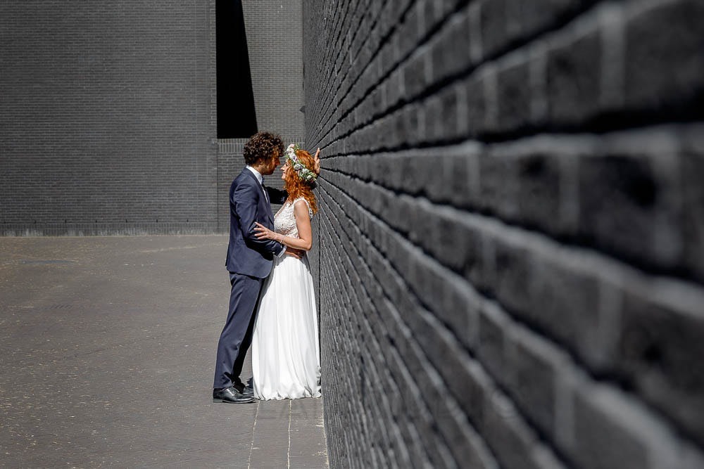 Para młoda, on w granatowym garniturze, ona w białej sukni z koronkową górą i wiankiem z kwiatów, opiera się o ciemnoszarą ceglaną ścianę, delikatnie się obejmując.