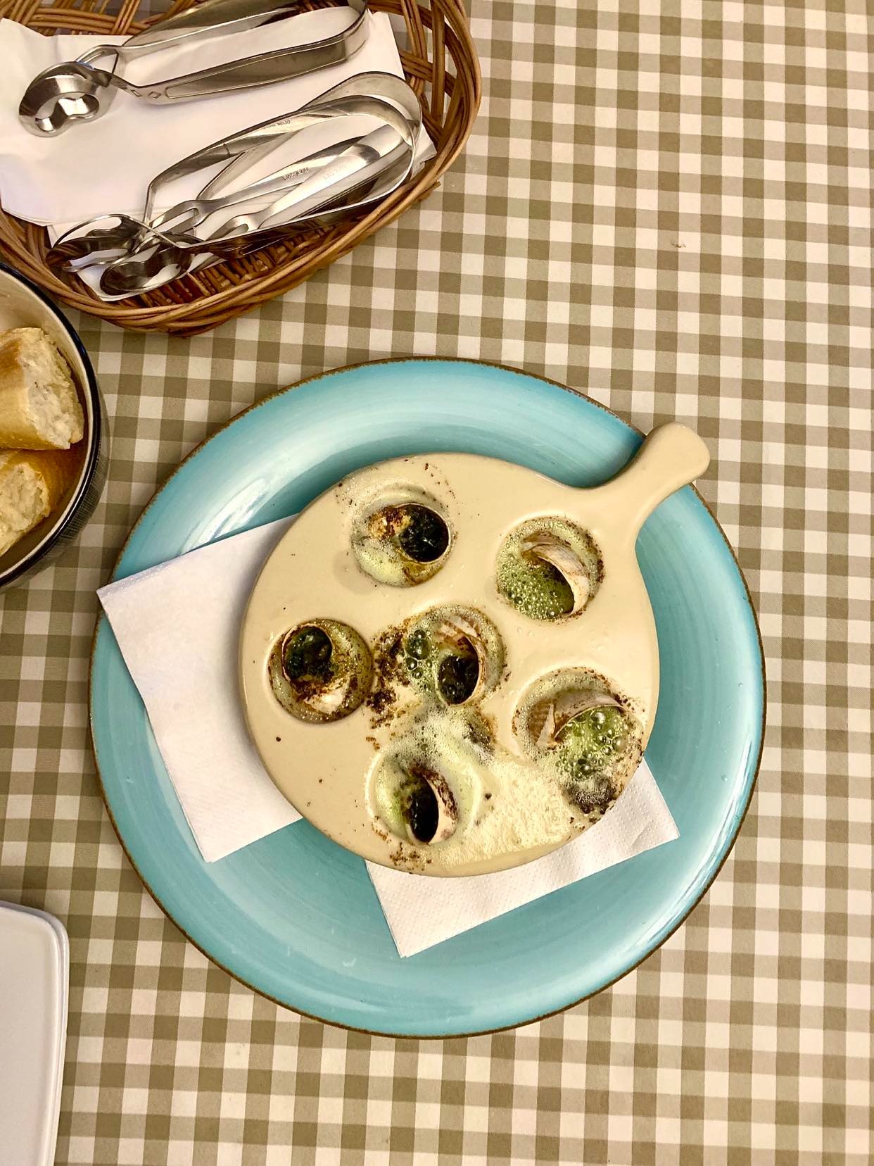 Escargots a la bourguignonne podane na niebieskim talerzu z chlebem i szczypcami do ślimaków, widok z góry na stole w kratę.