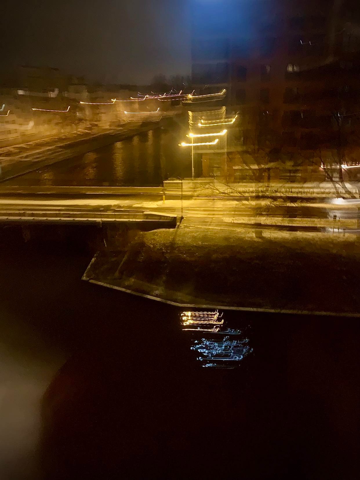 Nocny widok na kanał z odbijającymi się światłami lamp, sfotografowany zza szyby, tworzący efekt rozmycia i nakładania się warstw obrazu.