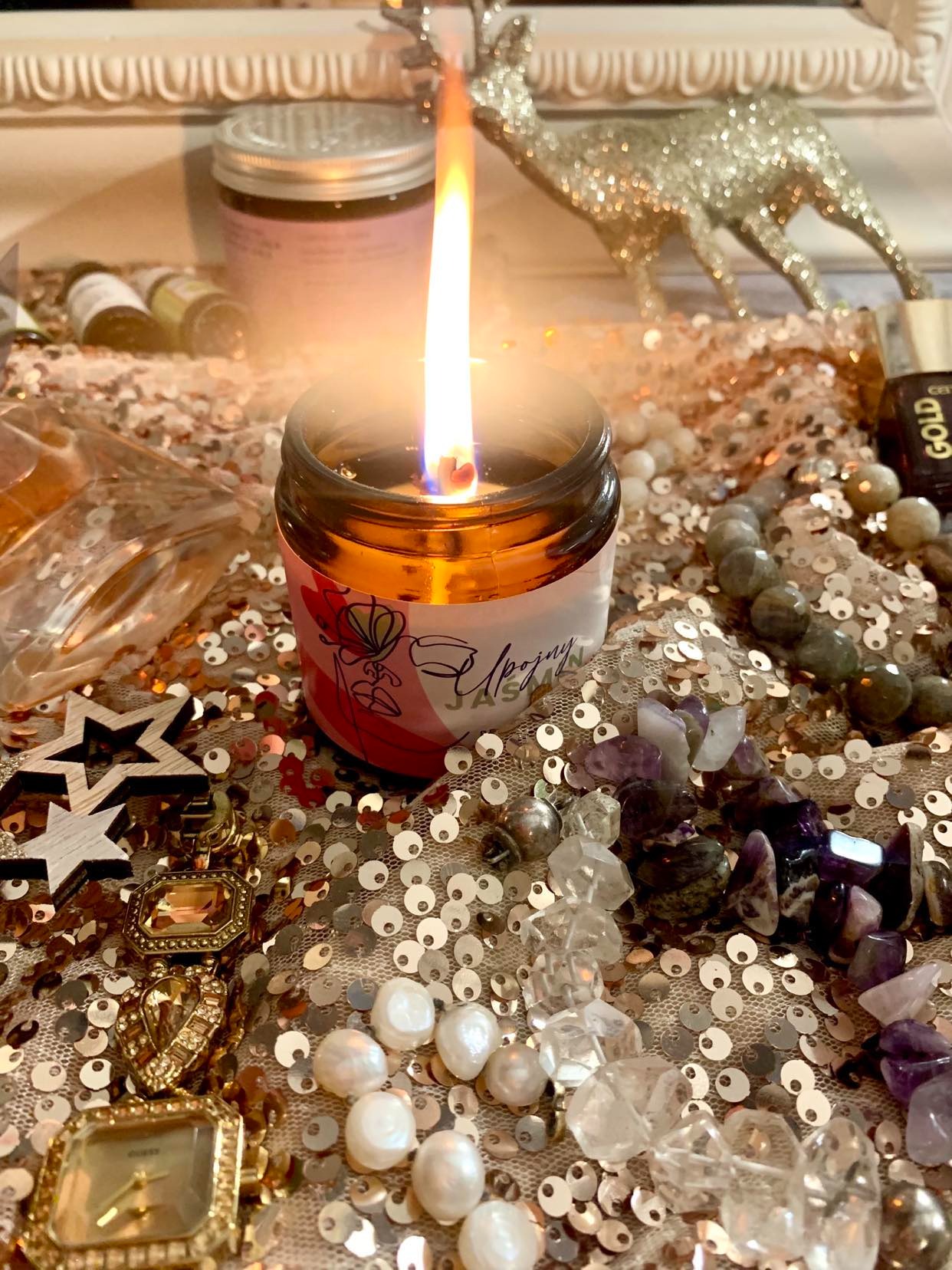 Płonąca świeca sojowa w szklanym słoiczku z etykietą kwiatową, otoczona biżuterią, kamieniami szlachetnymi, błyszczącymi cekinami i dekoracyjnym reniferem w tle.