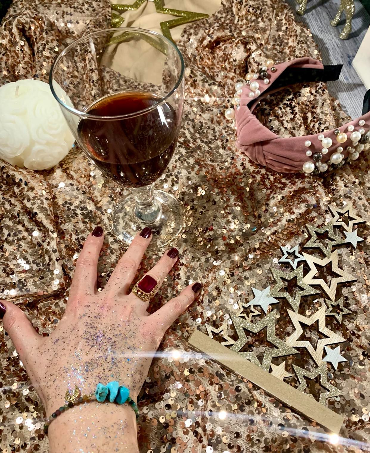 Kieliszek czerwonego wina na złotym, cekinowym tle obok ręki z brokatem, pierścionkiem z czerwonym kamieniem i bransoletką z turkusowymi kamieniami, różowa opaska z perłami, drewniane gwiazdki...