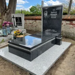 Granitowy nagrobek z czarną tablicą, na której widnieje portret zmarłego i inskrypcja, otoczony kwiatami i zniczami, na tle ceglanego muru i drzew