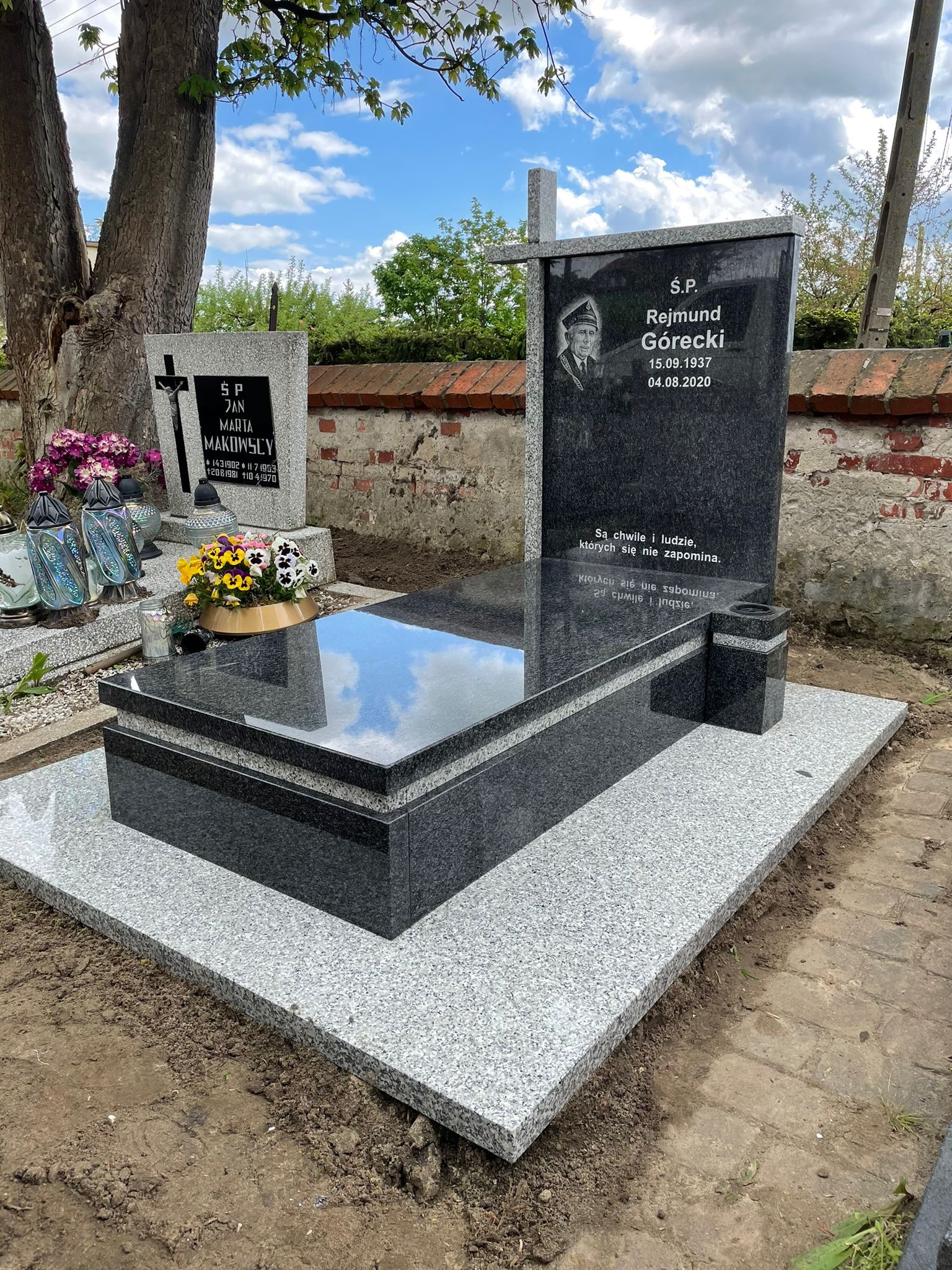 Granitowy nagrobek z czarną tablicą, na której widnieje portret zmarłego i inskrypcja, otoczony kwiatami i zniczami, na tle ceglanego muru i drzew