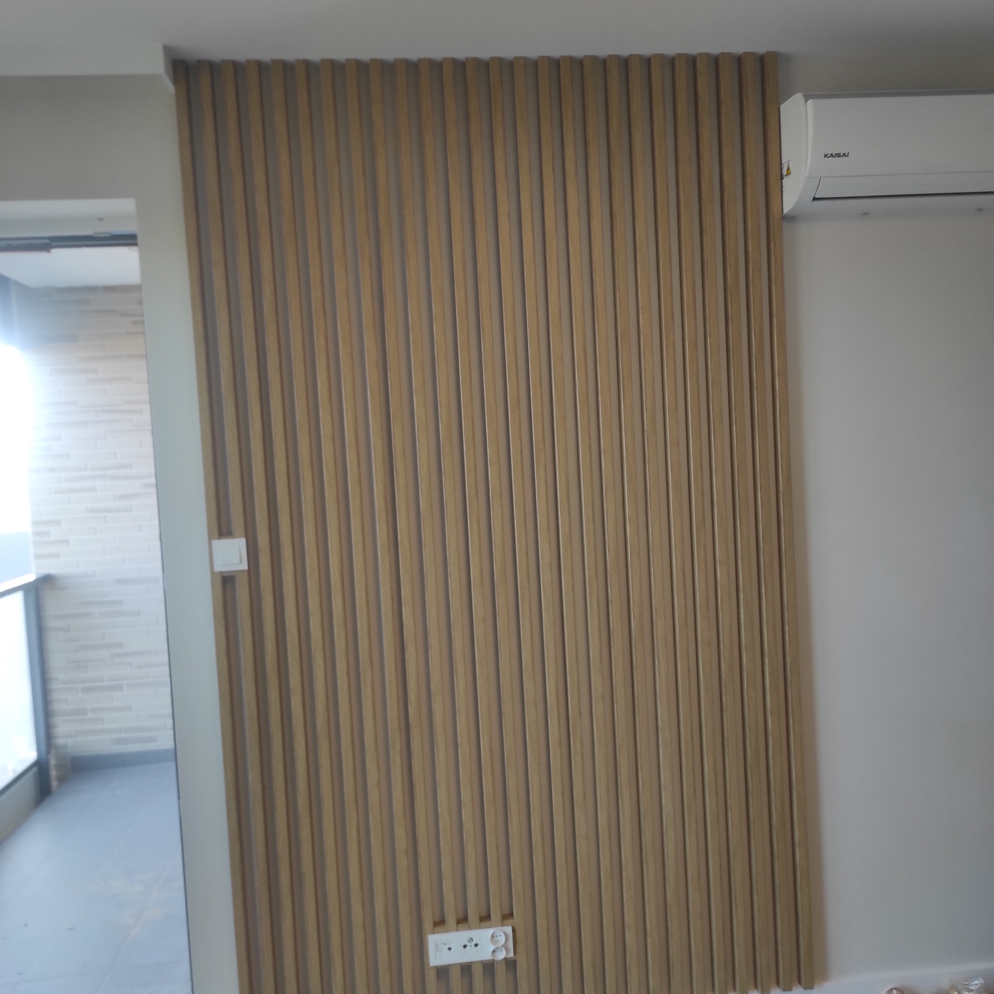 Drewniane lamele dekoracyjne na ścianie w jasnym odcieniu drewna, z widocznym gniazdkiem elektrycznym i klimatyzatorem Kaisai, tworzące nowoczesny wystrój wnętrza.