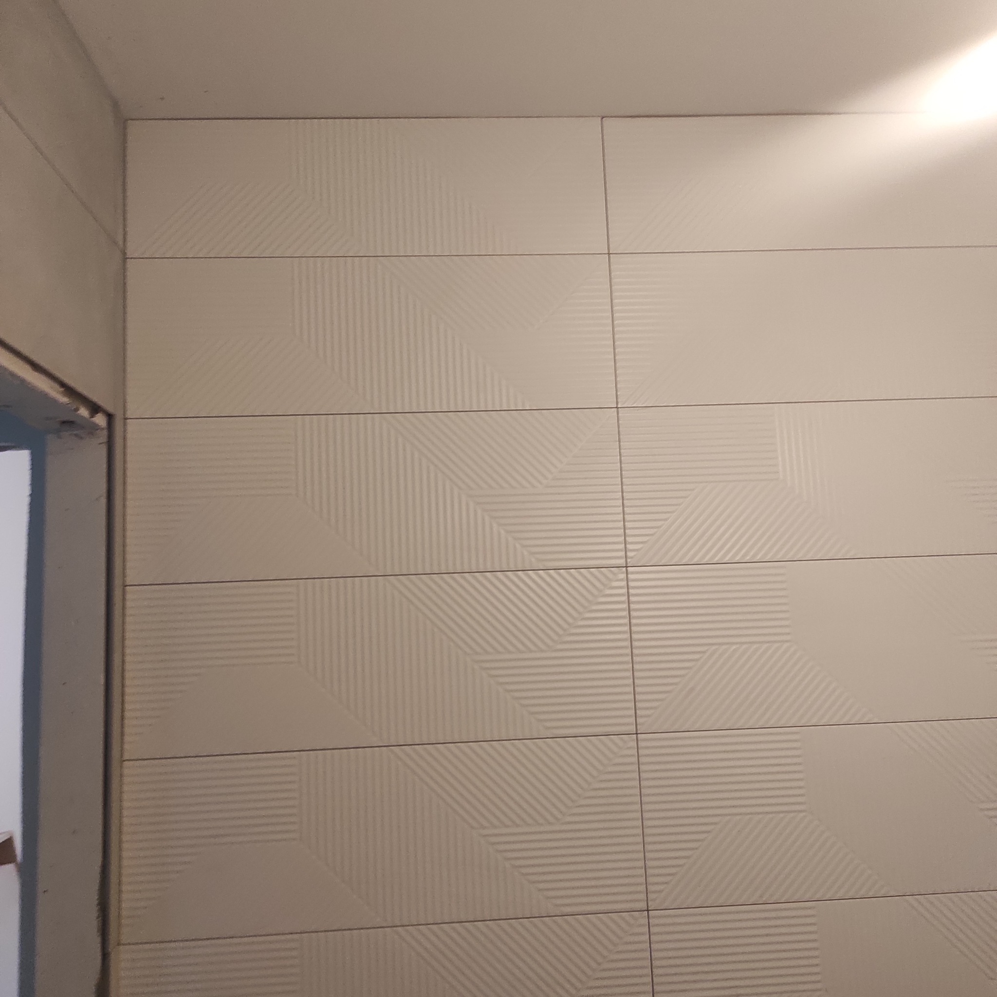 Ściana wyłożona beżowymi płytkami z delikatnym geometrycznym wzorem, widoczny fragment futryny drzwi.