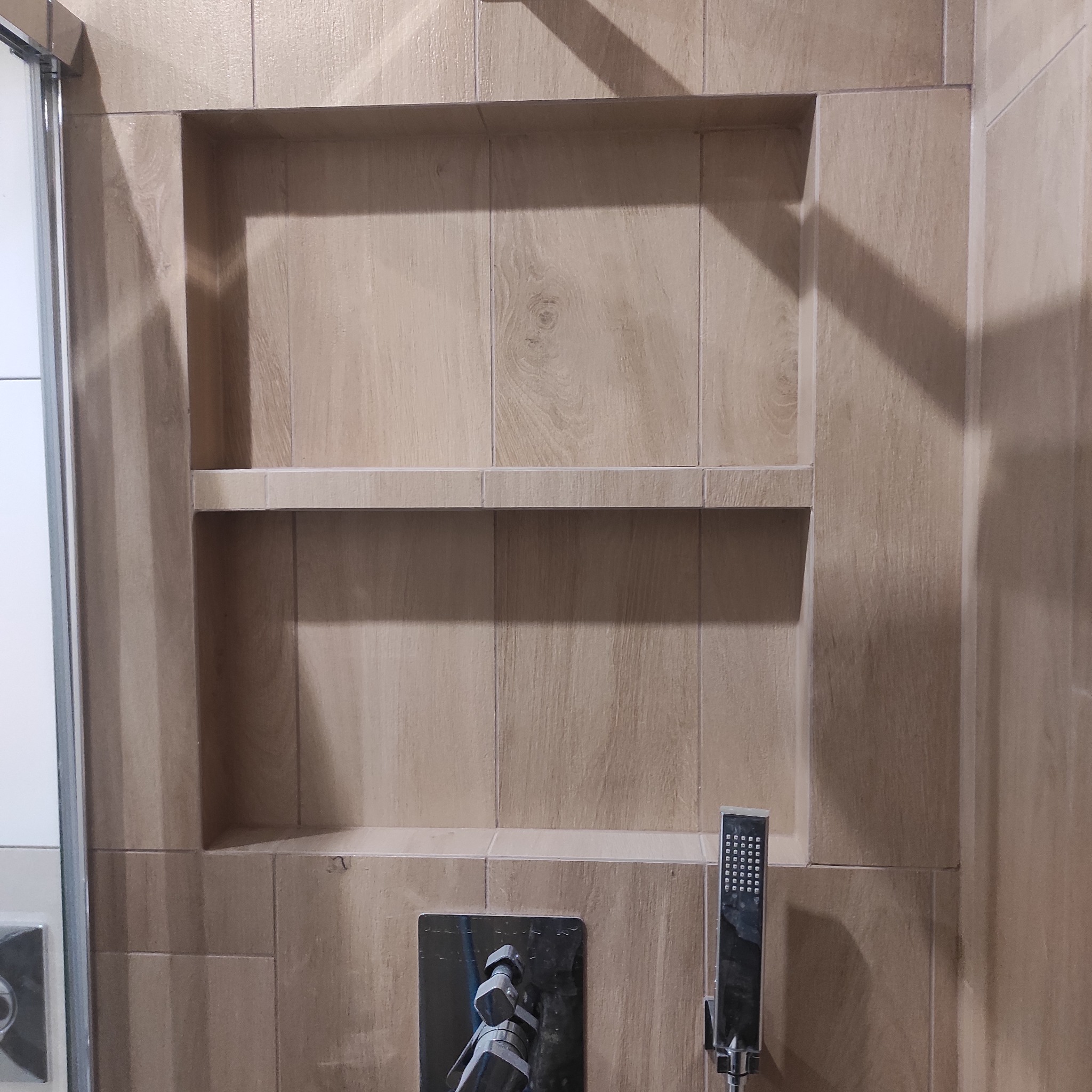 Narożna półka w kabinie prysznicowej wyłożonej płytkami imitującymi drewno, z widoczną baterią i słuchawką prysznicową.