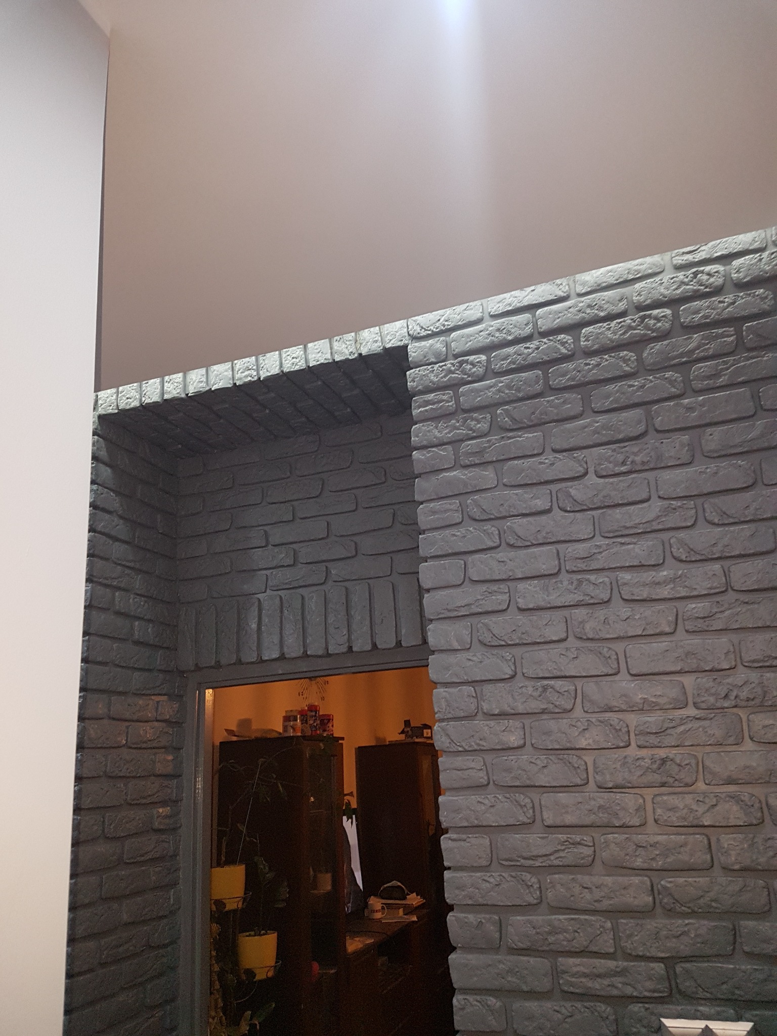 Szara ściana z cegły dekoracyjnej tworząca obramowanie otworu drzwiowego, widok na wnętrze pokoju z ciemnymi meblami i roślinami w doniczkach.