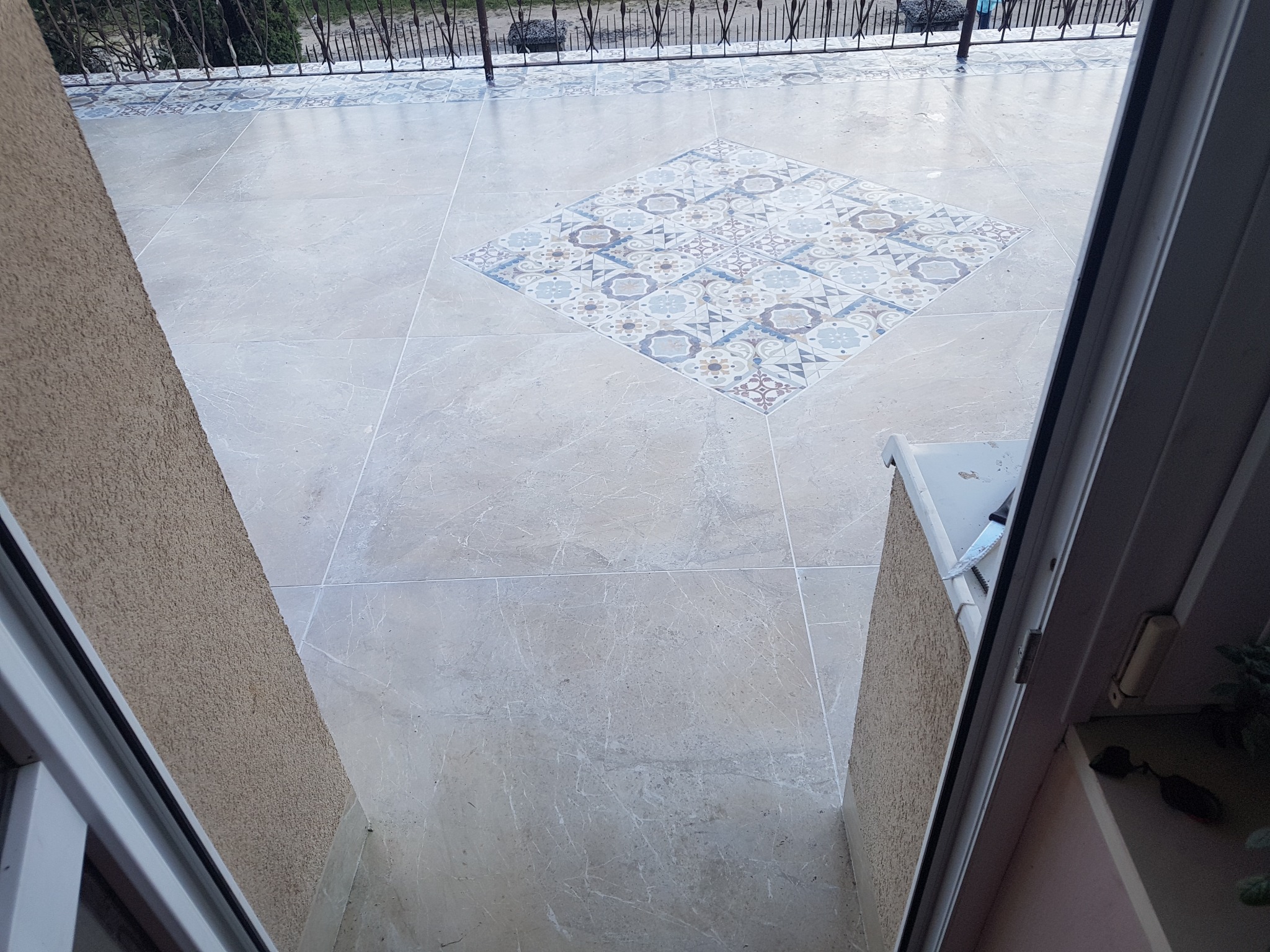 Wyłożony jasnymi płytkami taras z dekoracyjnym wzorem w centralnej części, widok z wnętrza przez otwarte drzwi balkonowe.