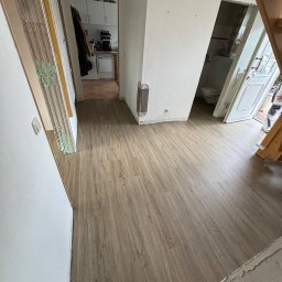 Daniel Obszyński - Korytarz z nową podłogą w odcieniach brązu, widoczne ślady po remoncie na ścianach i framugach, drzwi do łazienki i kuchni w tle.