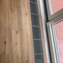 Daniel Obszyński - Jasnobrązowa podłoga z paneli, z wbudowaną metalową kratką wentylacyjną przy oknie. Widoczne niedoskonałości przy łączeniu paneli i ściany.