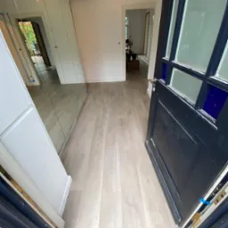 Jasna podłoga w korytarzu widziana zza otwartych, ciemnych drzwi z szybami w niebieskiej ramie, po lewej stronie lustrzana szafa.