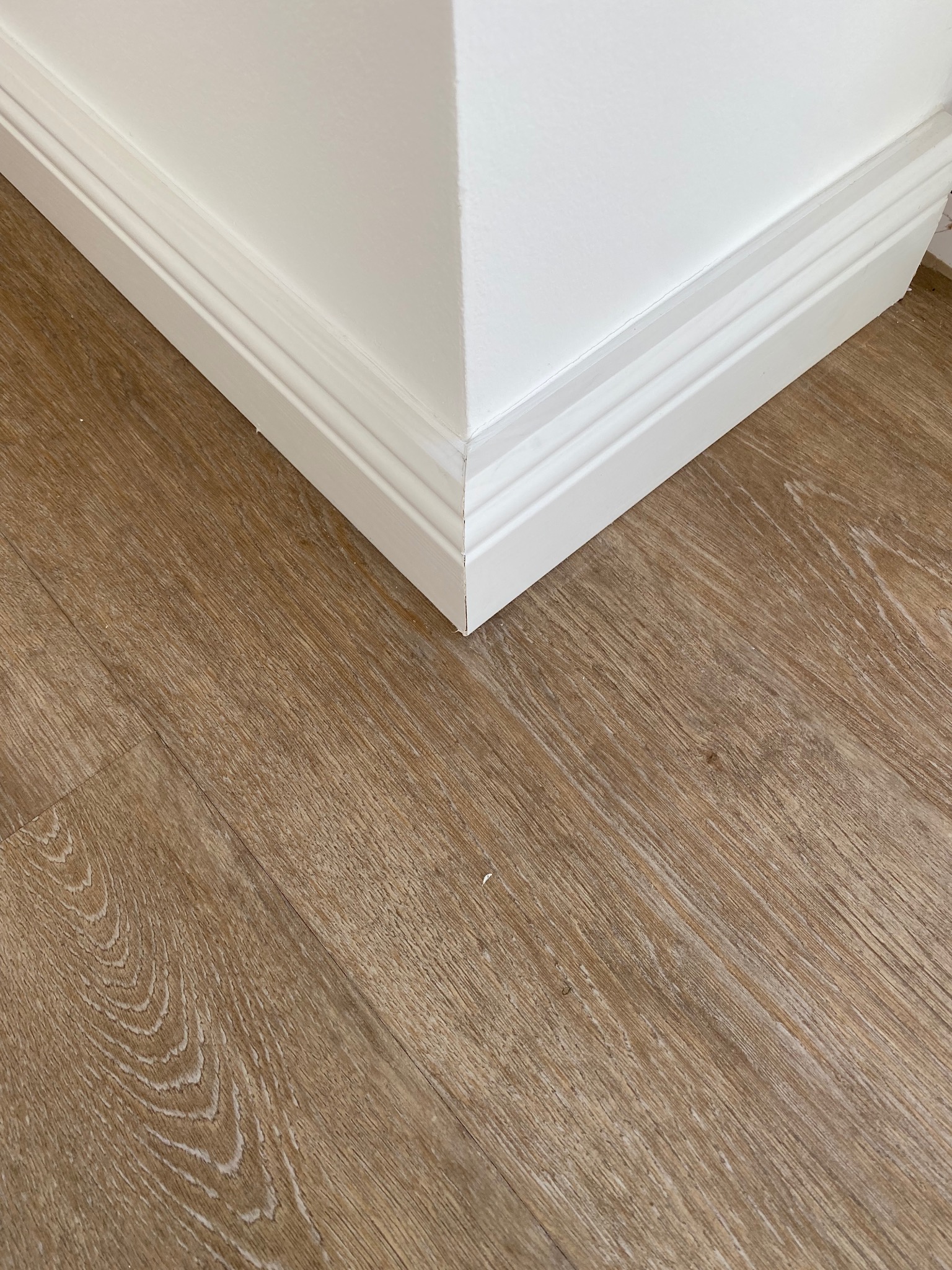 Jasne panele podłogowe w odcieniach drewna, ułożone przy białej listwie przypodłogowej w rogu pomieszczenia.