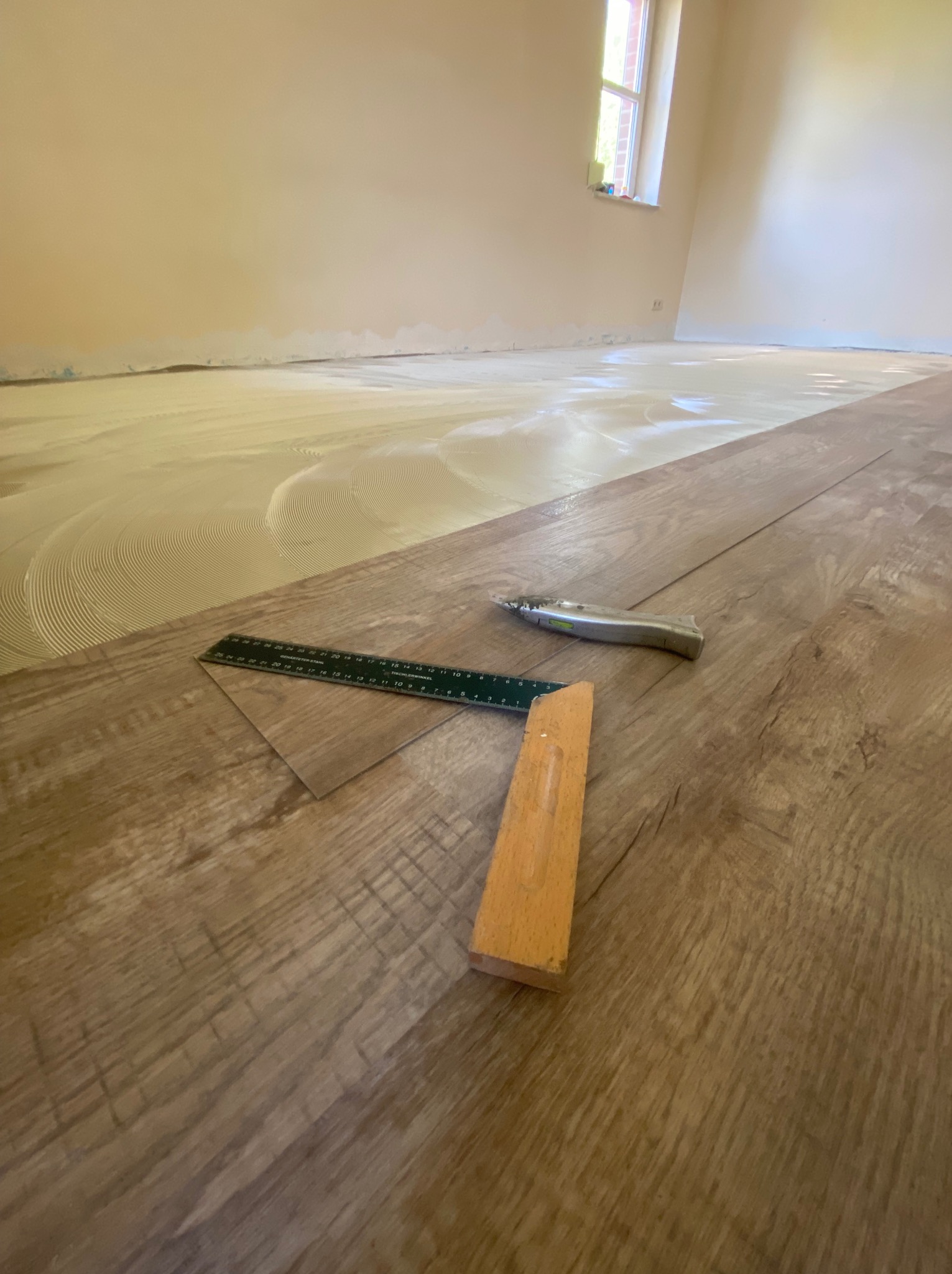 Fragment pokoju w trakcie prac wykończeniowych, z widocznym klejem na podłodze, częściowo ułożonymi panelami, kątownikiem, miarką i nożem monterskim.