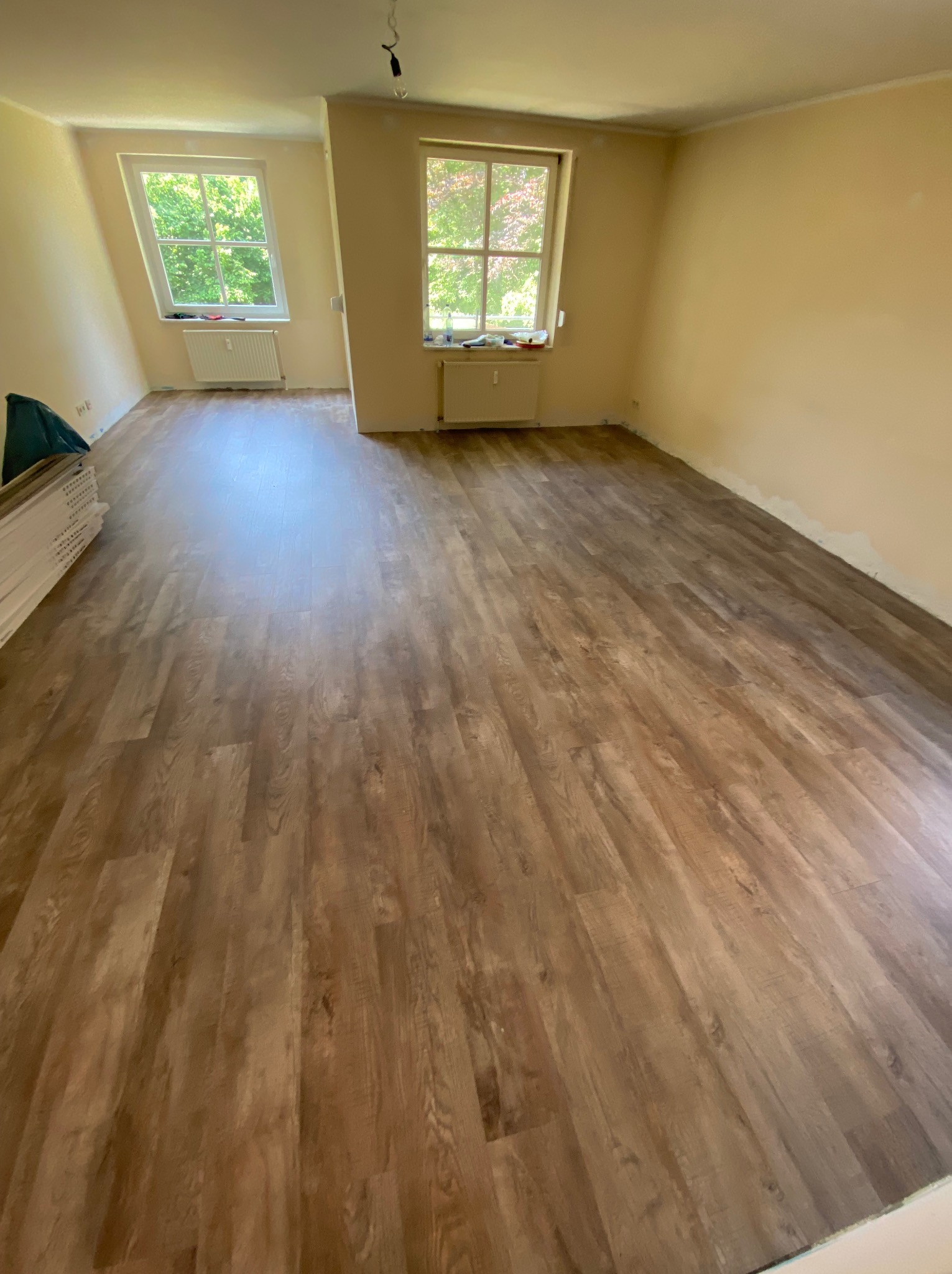 Pokój po remoncie z nową, brązową podłogą z paneli. W rogu pokoju leżą jeszcze nieużyte panele. Dwa okna wpuszczają naturalne światło.