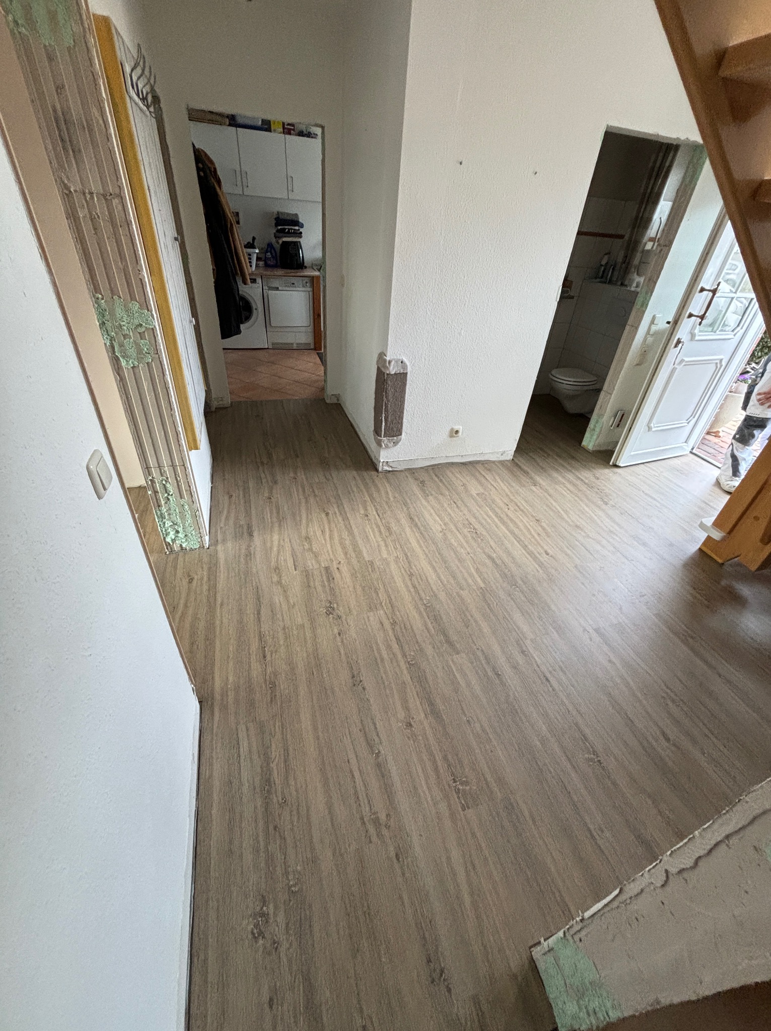 Korytarz z nową podłogą w odcieniach brązu, widoczne ślady po remoncie na ścianach i framugach, drzwi do łazienki i kuchni w tle.