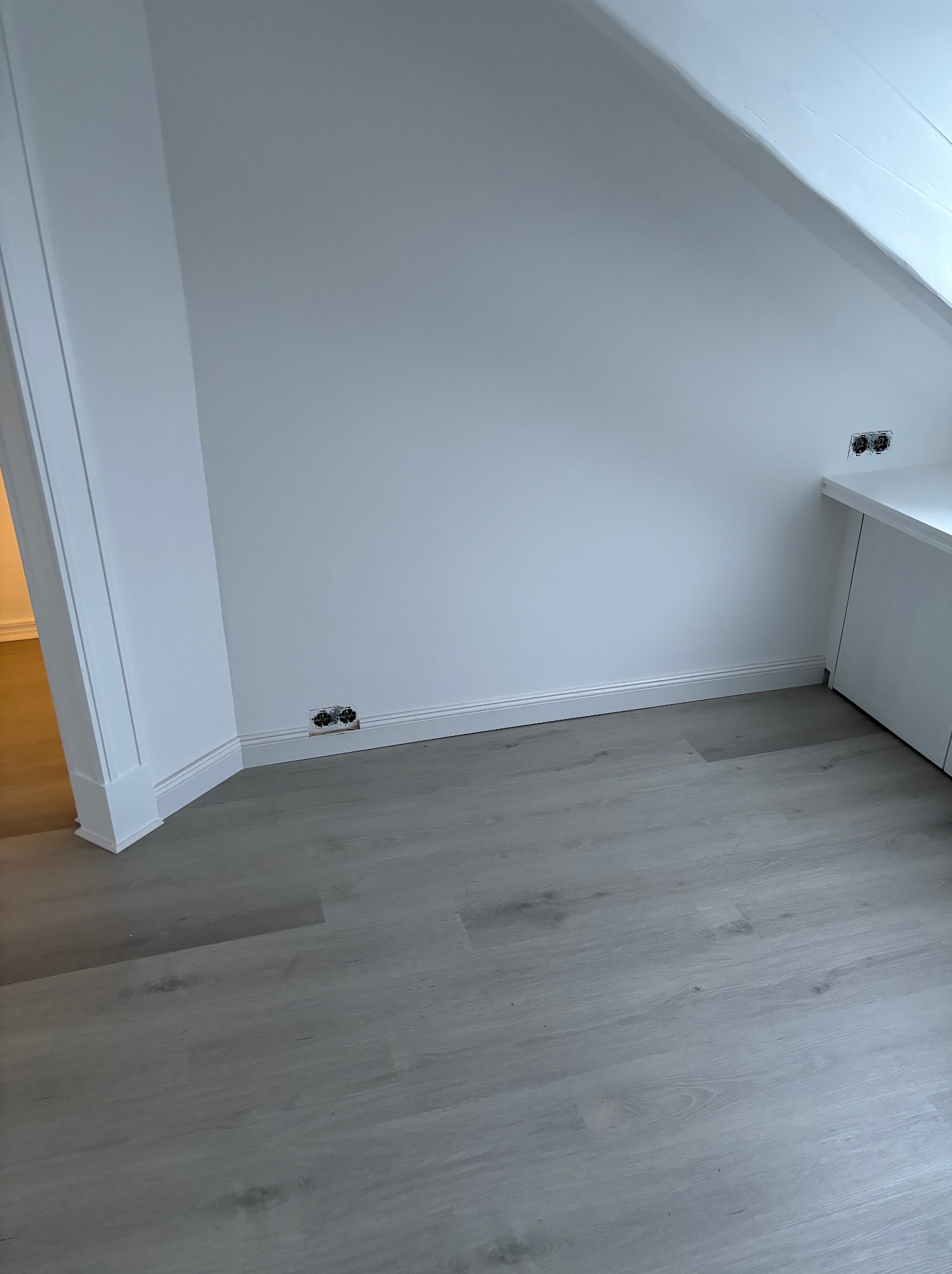Jasnoszare panele podłogowe ułożone w pokoju ze skosami, widoczne gniazdka elektryczne przy podłodze i białe listwy przypodłogowe.