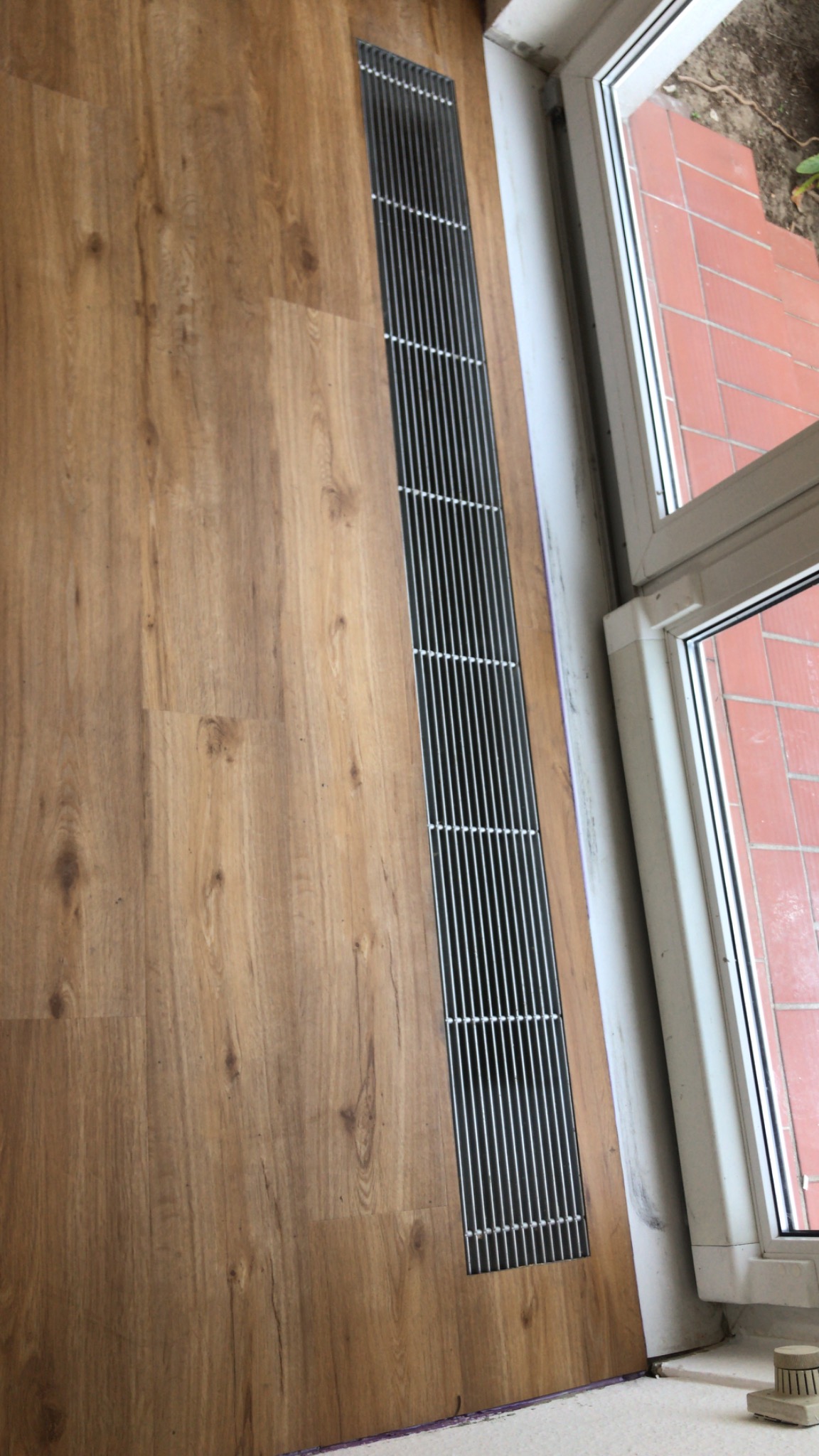 Jasnobrązowa podłoga z paneli, z wbudowaną metalową kratką wentylacyjną przy oknie. Widoczne niedoskonałości przy łączeniu paneli i ściany.