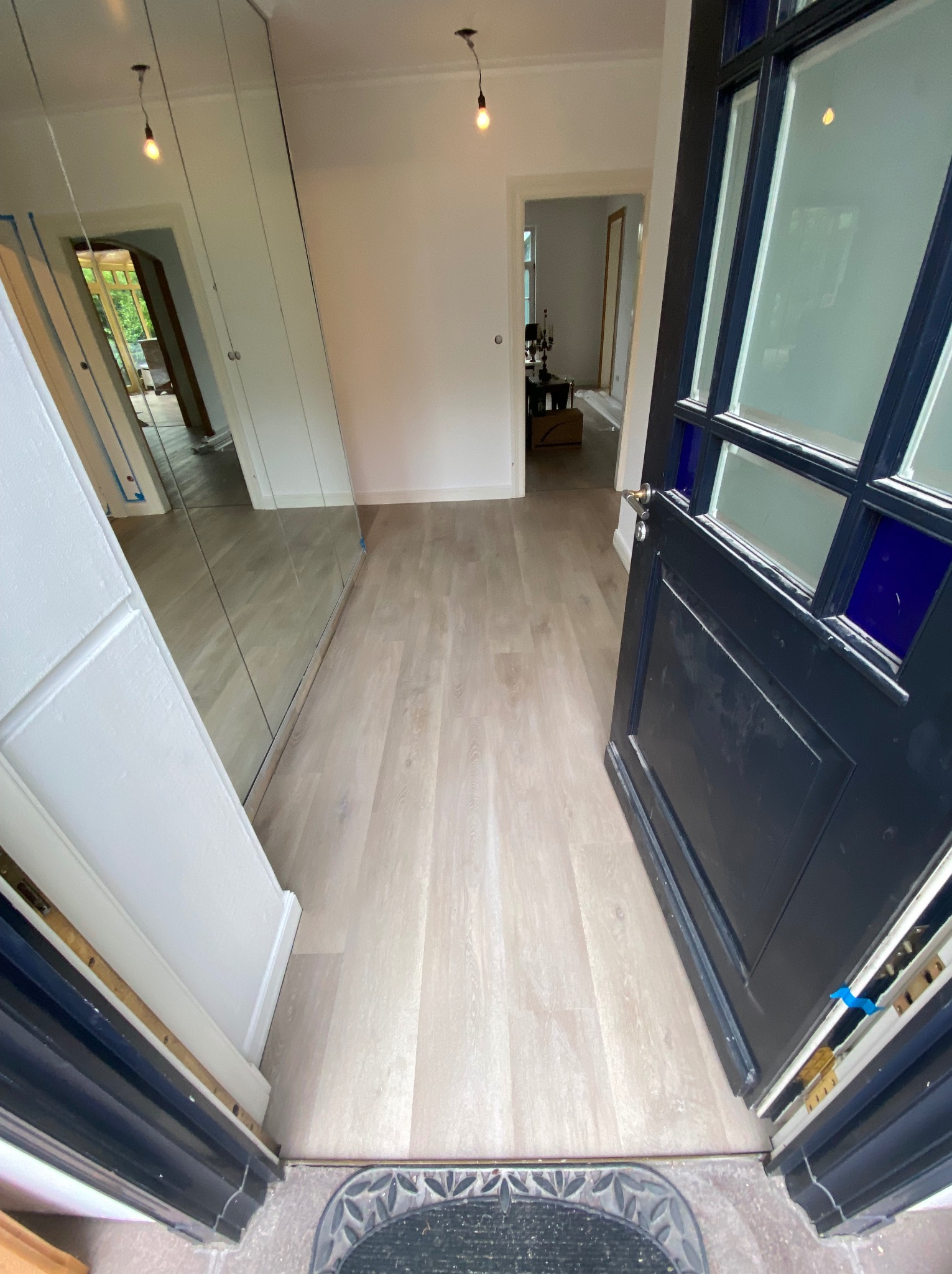 Jasna podłoga w korytarzu widziana zza otwartych, ciemnych drzwi z szybami w niebieskiej ramie, po lewej stronie lustrzana szafa.