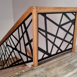 NORIX GM SPÓŁKA Z OGRANICZONĄ ODPOWIEDZIALNOŚCIĄ - Nowoczesna balustrada schodowa z jasnego drewna z czarnym, geometrycznym wypełnieniem, widoczna z góry na tle białych ścian i drewnopodobnej podłogi.